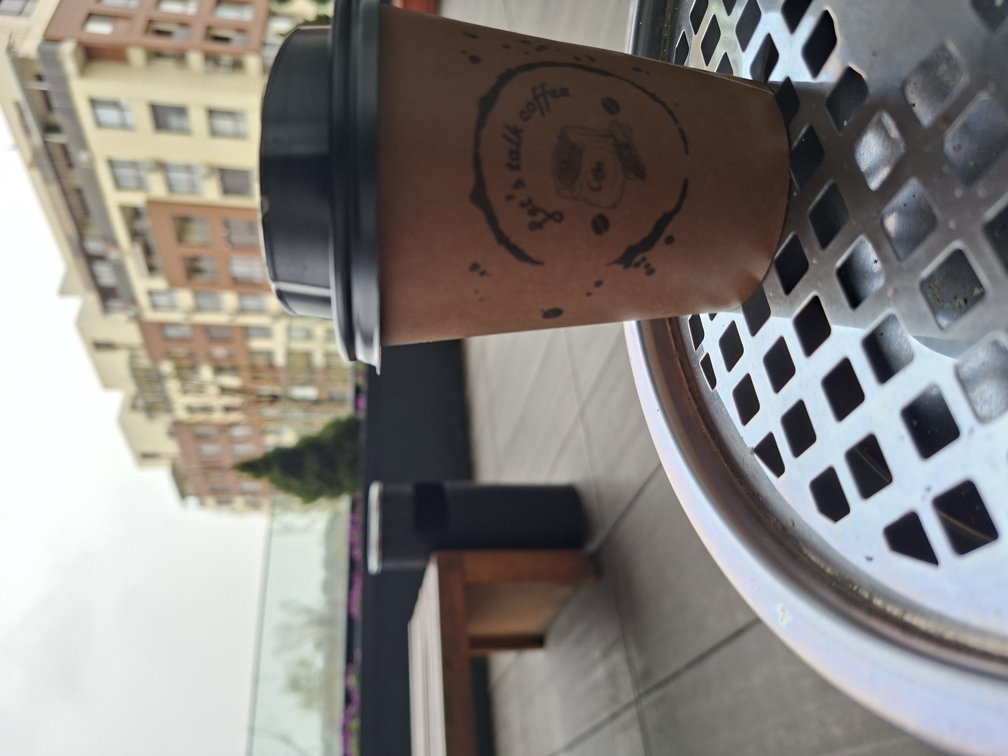 The image shows a coffee cup with a black lid resting on a metal trash can. The cup has a logo that says "Let's Talk Coffee". The background reveals an urban setting with a multi-story building, a glass barrier, and a bench. The location is Plovdiv, Bulgaria.