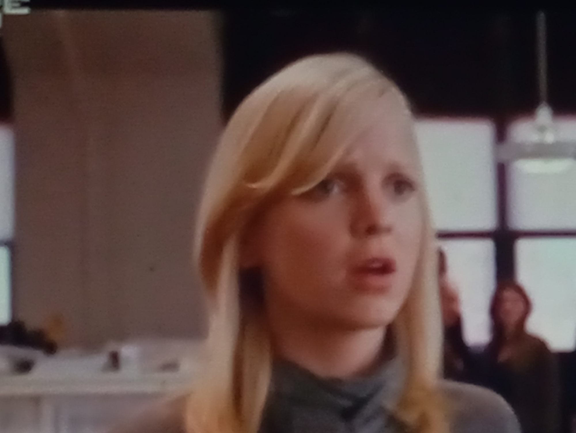 The media file is a close-up shot of a young woman with blonde hair styled in a layered cut. Her expression is one of surprise or concern, with her mouth slightly open and eyes wide. She is wearing a dark-colored, possibly gray, collared shirt. The background is blurred, suggesting an indoor setting. Behind her, several other figures are visible, also out of focus, and some appear to be looking in the same general direction as the main subject. In the far background, there are indications of windows or partitions that divide the space. The lighting suggests it could be daytime, as the background appears bright through the windows. There is no visible text in the image. The overall impression is a candid or dramatic moment captured from a film or video.