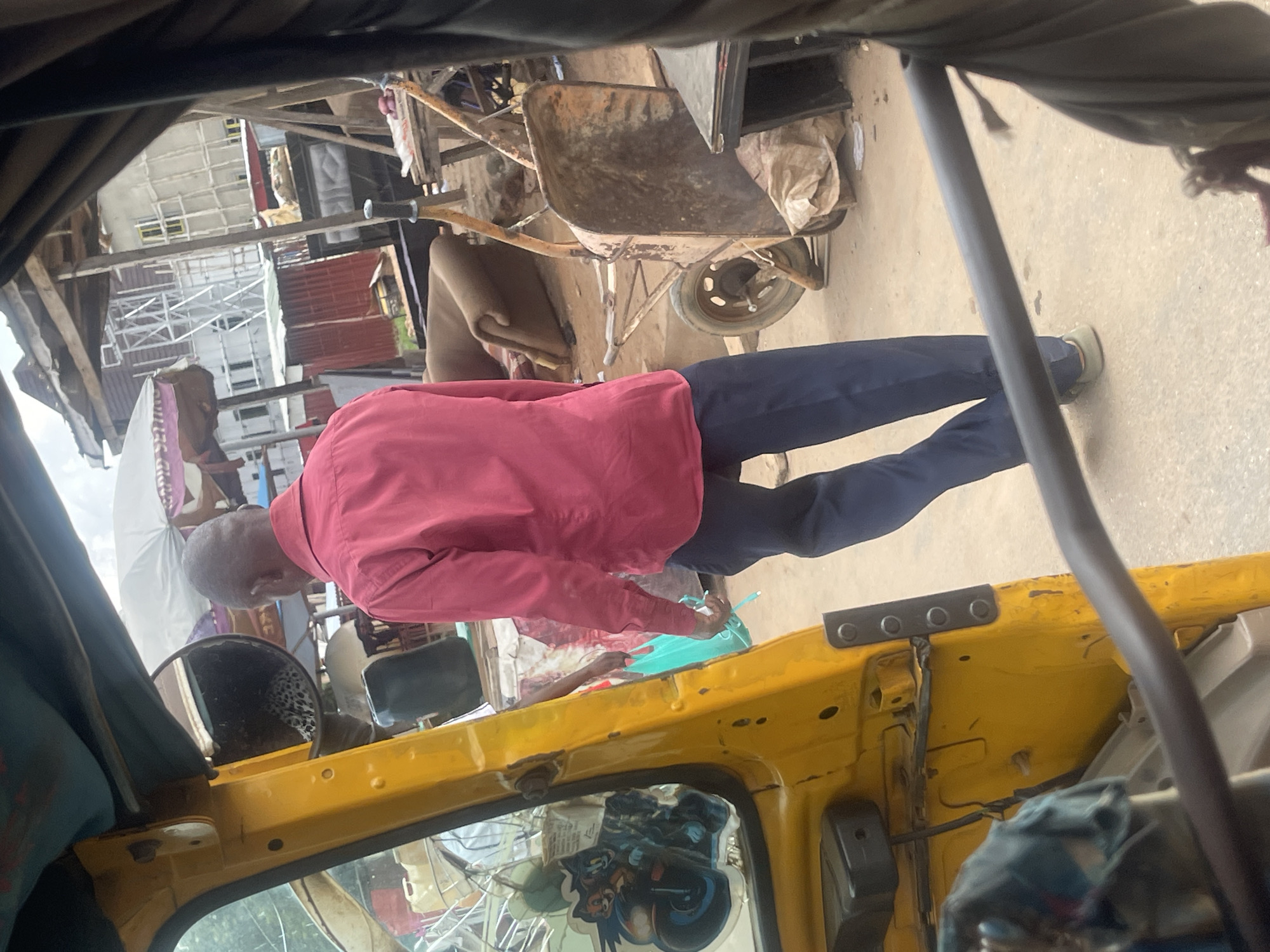 The image depicts an outdoor scene, likely in an urban environment, given the presence of a yellow auto-rickshaw, a wheelbarrow, a sofa, and buildings under construction in the background. A man in a red shirt and dark trousers is walking away from the camera, holding a light blue plastic bag. He is positioned to the right of the auto-rickshaw. The auto-rickshaw has a reflective mirror on its side, showing a leopard-print fabric inside. There are stickers on the window of the auto-rickshaw. The ground appears to be paved. The lighting suggests it is daytime. The overall impression is that of a busy, possibly developing area within a city.
