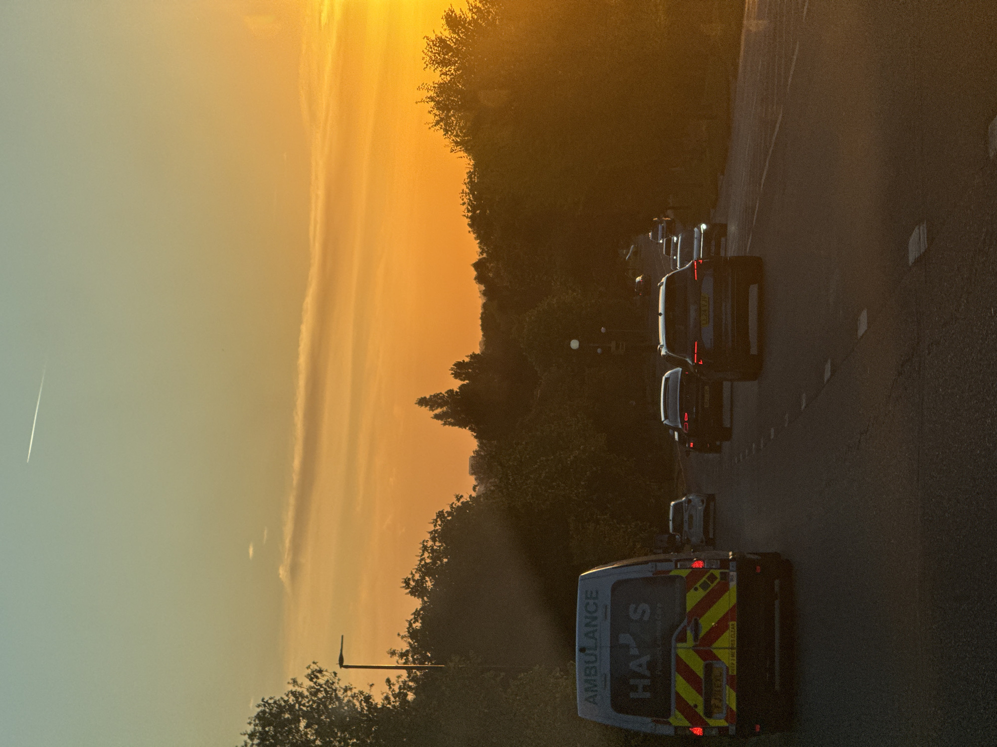 The image depicts a road scene during either sunrise or sunset, characterized by a dominant warm, golden light. The perspective appears to be from inside a vehicle, looking forward at the road and surrounding environment.
In the immediate foreground, the rear of a white ambulance is prominently featured. It displays distinctive yellow and red chevron markings, alongside illuminated red taillights. Visible text on the ambulance includes "AMBULANCE" written vertically on the left side, and large letters "HA S" centrally. Below these, a partial message "Keep 1.5M clear" can be deciphered. The ambulance's license plate reads "LU21 LSY".
Ahead of the ambulance, several other vehicles, including cars and vans, are visible on the road, forming a line of traffic. The license plate "LX21 LCV" is discernible on a white car further up the road. The road surface itself shows lane markings and appears to be a multi-lane highway or main road.
To the right side of the road, a dense line of trees and foliage forms a dark silhouette against the bright sky, accentuated by the low-angle sun. A street light pole is also visible on the left, near the treeline.
The sky, filling the upper and left sections of the frame, displays a striking gradient of colors. It transitions from an intense orange and yellow where the sun's light is strongest, to a softer, lighter blue-grey further away. Several wispy clouds stretch across the sky, illuminated by the golden hues. A faint, white contrail from an aircraft is also visible in the upper left corner of the sky. The overall lighting suggests the tranquil ambiance of the "golden hour."
