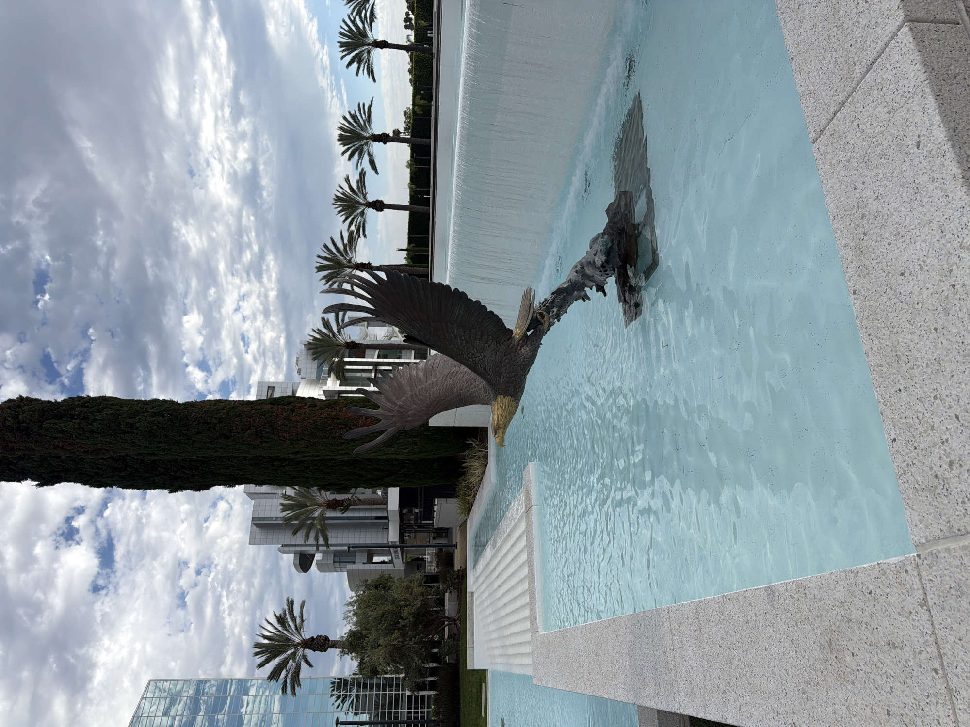 A statue of a bald eagle in mid-flight is prominently featured in a fountain. The eagle statue, made of dark metal, is positioned in the foreground, with its wings spread wide and its beak pointing forward. It appears to be emerging from or diving towards a dark, rocky base submerged in the fountain's water.
The fountain itself is multi-tiered, with the eagle statue in the lower section. The water in this section is a clear, light blue. Behind the eagle, a wide, white waterfall cascades down a smooth, vertical surface, creating a wall of water. The area surrounding the fountain is paved with light-colored stone, with some sections featuring tiered, stepped designs.
In the background, a stark contrast is presented by modern architecture. A tall, slender, dark green tree stands to the left of the waterfall. Further left, a tall, glass-facaded building with a blue reflective exterior dominates the skyline. Other modern, white buildings with curved elements and geometric shapes are visible behind the fountain and palm trees. Several palm trees are spaced out across the midground, their fronds reaching upwards. The sky above is partly cloudy, with patches of blue sky visible between formations of white and gray clouds. The scene appears to be an outdoor public space, likely in a city. This specific location is a public art installation at the Crystal Cathedral in Garden Grove, California, United States of America.