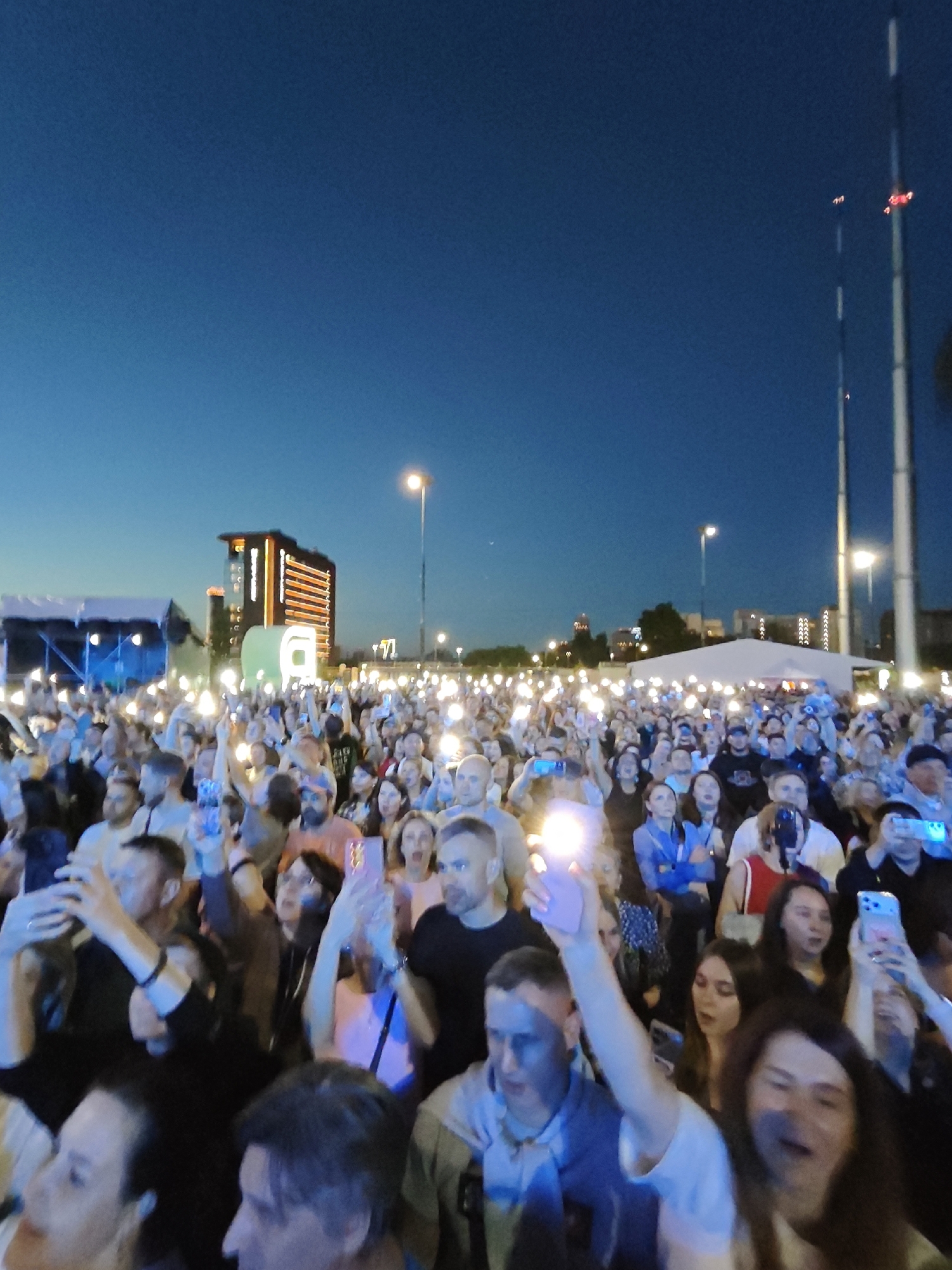 The image depicts a large crowd of people gathered outdoors at dusk, likely at a concert or festival. Many individuals are holding up their mobile phones, with their screens illuminated, suggesting they are recording or photographing the event. In the background, a modern, illuminated building stands out, with a series of bright vertical orange lights on its facade. Other structures, including what appears to be a stage or canopy covered by a white tent, are visible. Tall, slender poles, possibly lighting towers or communication masts, are also present in the scene. The sky is a deep blue, indicating the transition from daylight to night. The overall atmosphere suggests an urban event with a significant audience. Based on the architectural style of the illuminated building and its context within a cityscape, this scene is likely located in Yekaterinburg, Russia. The building appears to be the "Vysotsky" Business Center, a prominent landmark in Yekaterinburg.