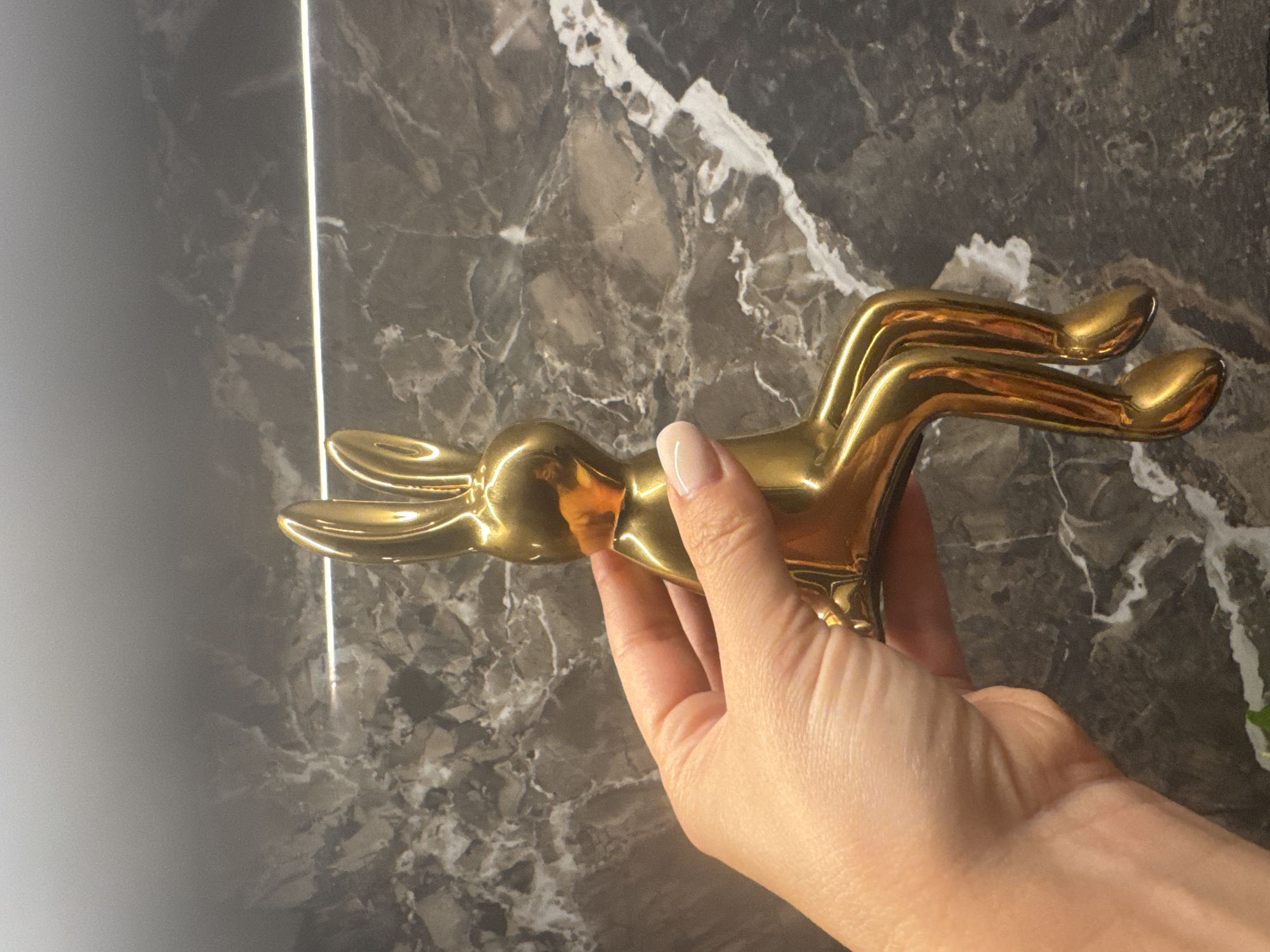 A person's hand with light-colored fingernails is holding a shiny, gold-colored rabbit figurine. The figurine is sitting with its legs extended and is oriented vertically in the frame. The background is a dark, marbled surface with white veining and a thin, bright horizontal line running across it. The image appears to have been taken indoors. The location in the city of Sochi, Russia is not discernible from the image.
