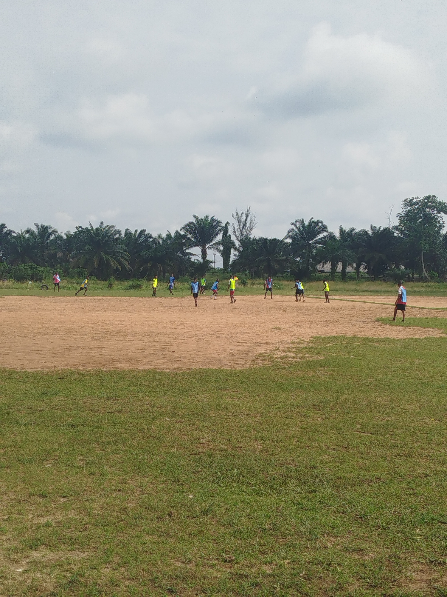 A soccer game is in progress on a dirt field. Players wearing yellow and blue jerseys are scattered across the field. A grassy area is in the foreground. The background consists of palm trees and cloudy skies. The location is Ikot Mbonde, Nigeria.