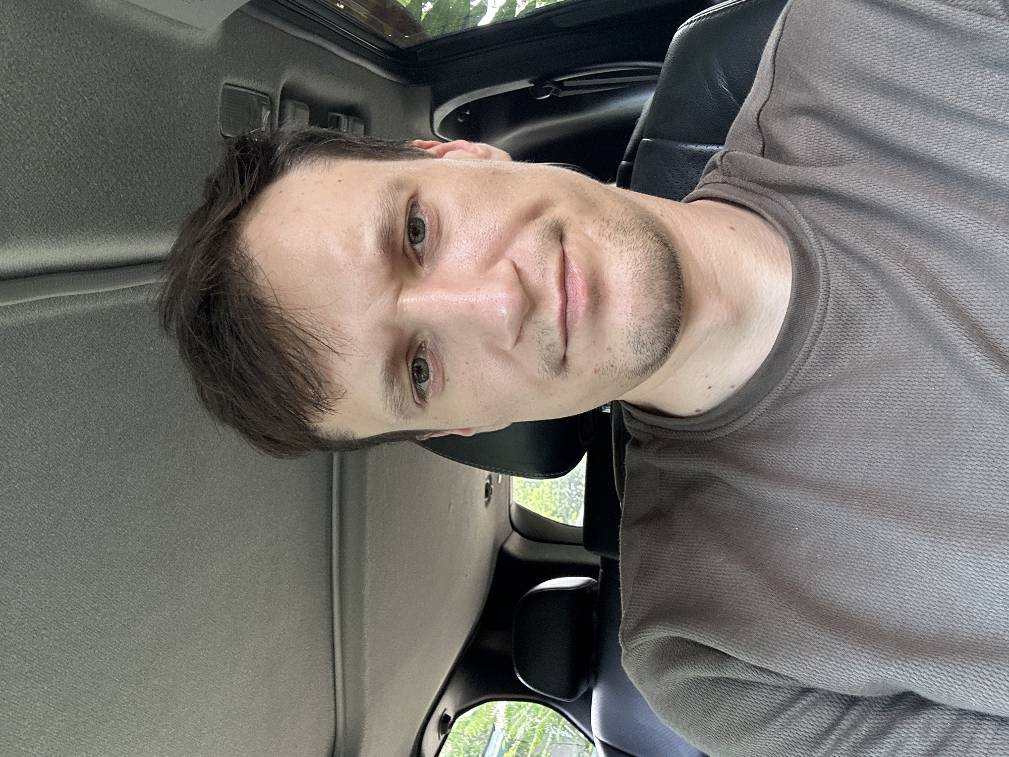 A man is pictured from the chest up, inside a vehicle, likely an SUV, given the visible headrests and interior trim. He is seated in the driver's seat, looking directly at the camera. He has short, dark brown hair and light-colored eyes. He is wearing a brown, textured long-sleeved shirt. The interior of the car is dark, with a gray headliner and black leather seats. Sunlight filters in from the windows, illuminating the scene. Outside the car, greenery and trees are visible, suggesting a natural or suburban environment. The image was captured in Makiv, Ukraine.