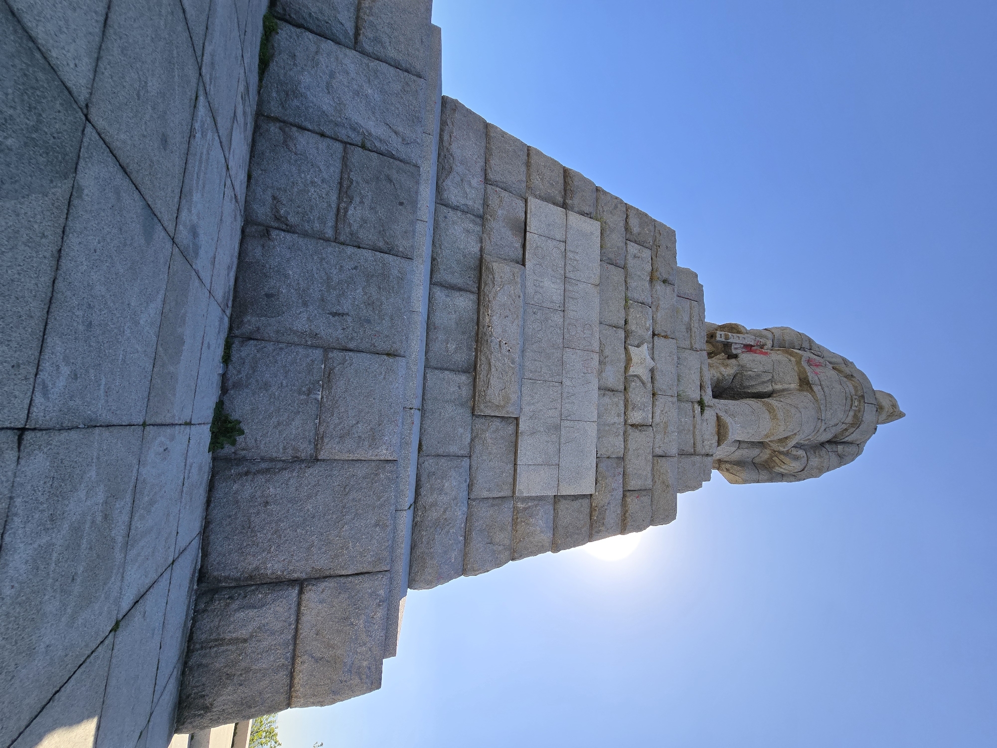 The image shows a statue against a clear blue sky. The statue is made of light grey stone and appears to be a figure in military attire. It stands atop a tiered stone structure made of large, rectangular blocks. The sun is behind the statue, creating a slight glare. The location is not specified, making the city and country unknown.