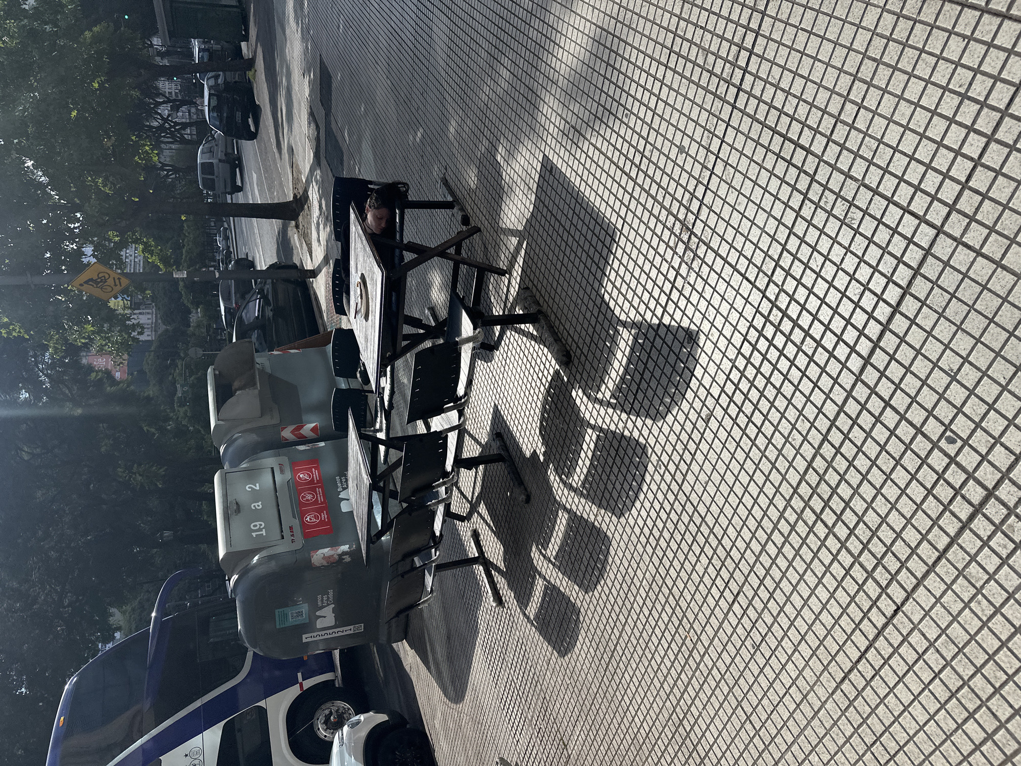 The image captures a street scene in Buenos Aires, Argentina. The foreground features a sidewalk paved with square tiles. A small outdoor cafe setup is visible, including a dark-colored table and chairs, casting shadows on the sidewalk. Adjacent to the cafe seating are large trash containers labeled "19 a 2" with Buenos Aires branding. A bus with a blue and white color scheme is parked beside the trash bins. In the background, across the street, are parked cars, trees, and buildings. A street sign depicting bicycles is visible. A woman is sitting at the table. The overall impression is that of a typical urban setting in Buenos Aires, with a blend of public transportation, street amenities, and outdoor commercial spaces.
