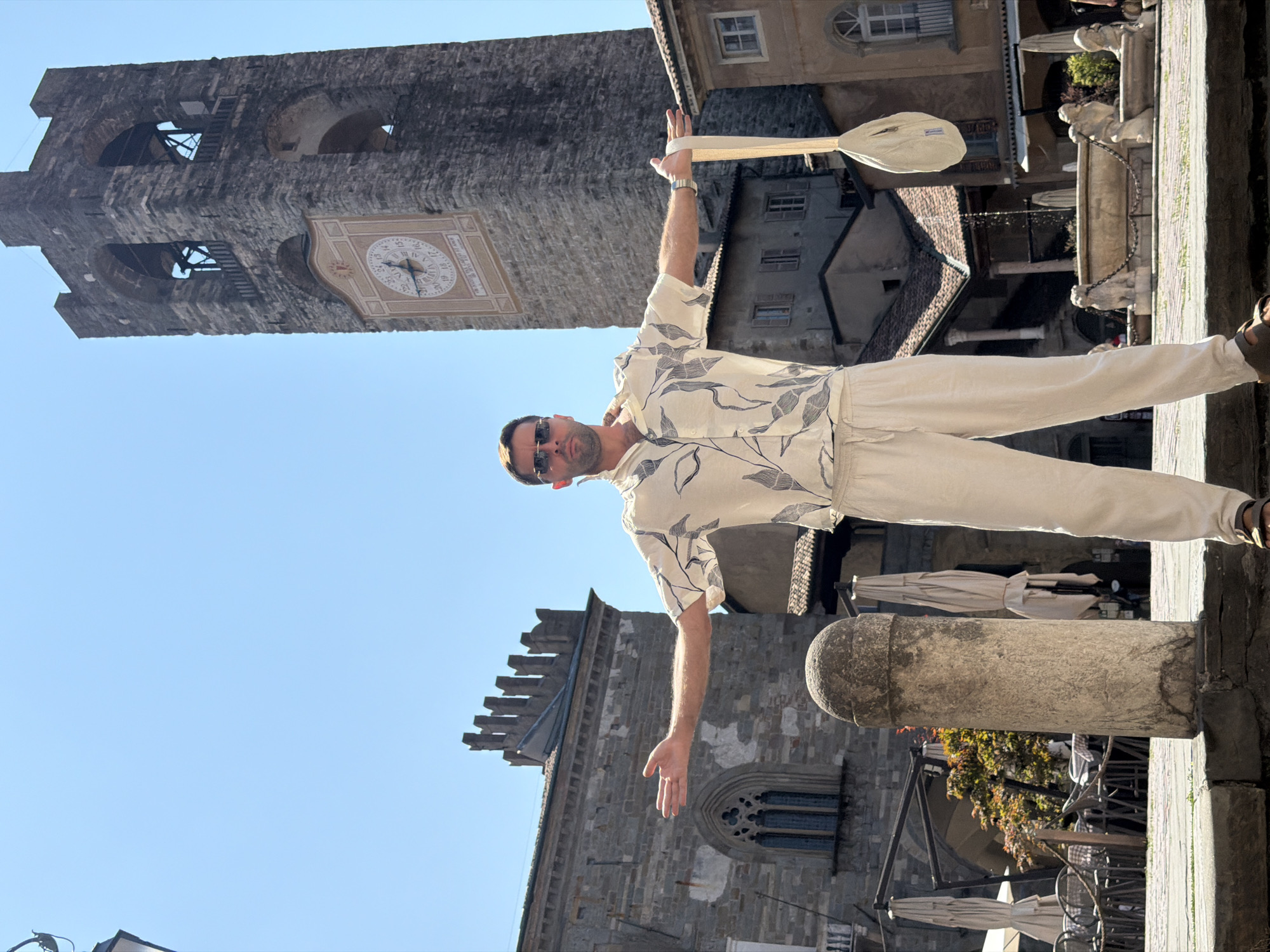 A man stands in a square in front of a tall stone clock tower with a clock face. He is wearing a light-colored short-sleeved button-down shirt with a dark leaf pattern and light-colored drawstring pants. The man is wearing sunglasses and has his arms outstretched to his sides. He is holding a light-colored bag by its strap in his right hand. The square has stone paving, a round stone pillar in the foreground, outdoor seating with umbrellas, and a fountain with lion statues in the background. The architecture visible suggests an Italian setting, possibly in the city of Mozzo, Italy. The sky is clear and blue.