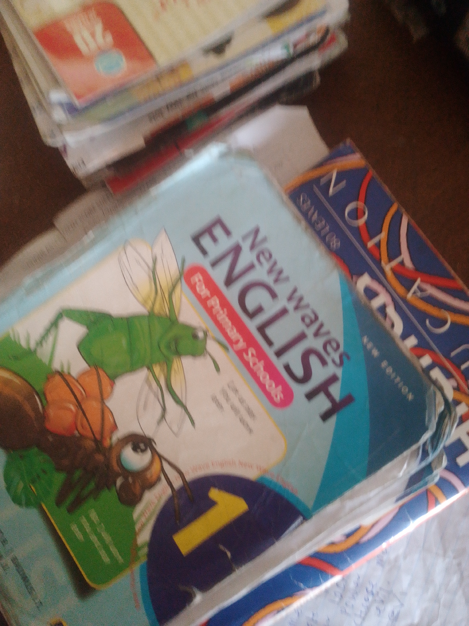 The image shows a stack of books. The top book has a blue cover with the words "New waves ENGLISH" in large, bold lettering. Below this, in a pink oval, it says "For Primary Schools". There is an illustration of an ant and a grasshopper on the cover. The book below this has a blue cover with a pattern of colorful, wavy lines and the letters "N O N C A". Other books are visible beneath these, with glimpses of text and illustrations. The scene appears to be indoors, possibly a classroom or a home, given the presence of books. The lighting suggests it is daytime. No specific time of day, weather, or emotional cues are discernible. The location context is Jauro Yinu, Nigeria, which provides information about the geographical setting. The visible text on the books provides details about their content, likely educational materials for primary school English.