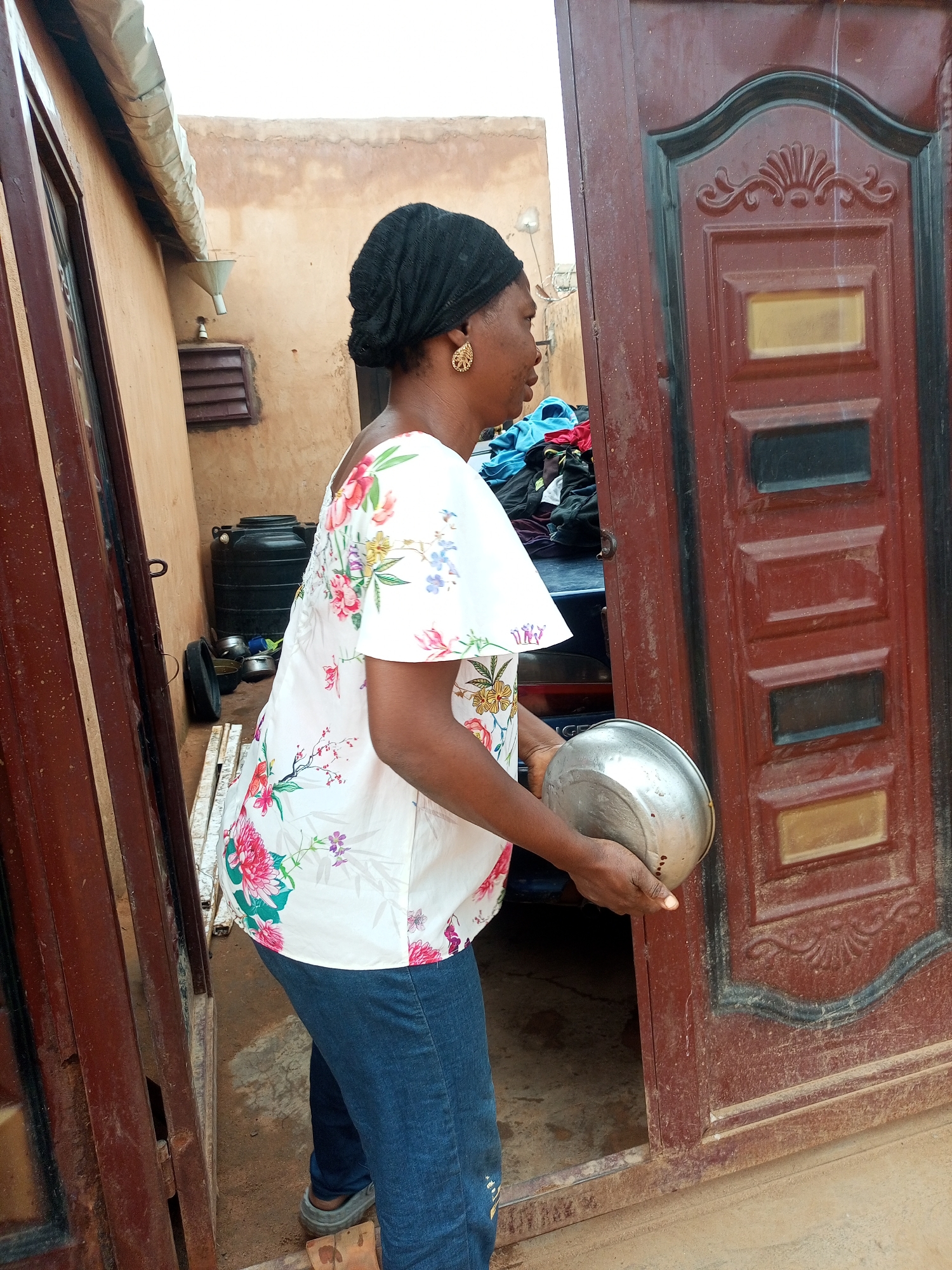 A woman wearing a black headscarf and a white shirt with a floral pattern stands in a doorway, holding a metal bowl. She is wearing blue jeans and grey shoes. She is facing to the left of the frame. Behind her, there is a brown door with ornate detailing, a black water tank, and a pile of clothes. The environment appears to be outdoors, possibly in a residential area.