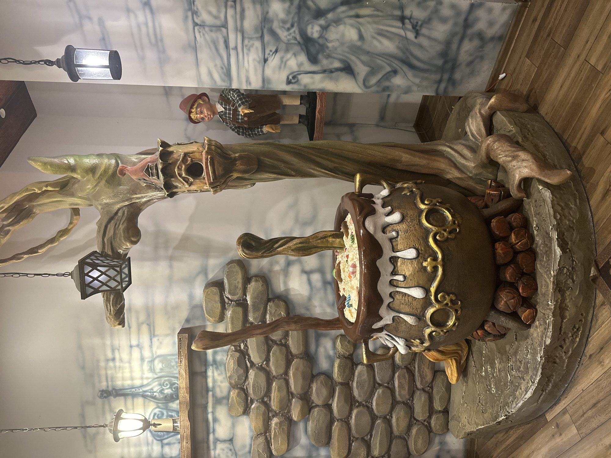 The image depicts a whimsical, theatrical display, likely an interior decoration. Dominating the foreground is a large, decorative cauldron filled with what appears to be candy and dough, resting on a base made to resemble rocks and logs. Two large wooden spoons are inserted into the cauldron's contents. Behind the cauldron, a stylized tree trunk sculpture rises, featuring a birdhouse with a small figurine of a bird or gnome on top. To the right of the tree, a small, static figurine of a young boy in overalls and a red hat stands on a shelf. The background features a stone-like wall texture and painted murals of a witch and bottles. Several hanging lanterns are visible, one on the left with a warm glow, and two on the right, one of which is also illuminated.
This scene is located at the "The House of Fairy Tales" (Къщата на Приказките) in Plovdiv, Bulgaria. The exhibit is designed to evoke a magical or storybook atmosphere, with each element contributing to a narrative theme. The lighting is atmospheric, with the lanterns providing soft illumination to highlight the display. The floor is made of wood-look laminate.