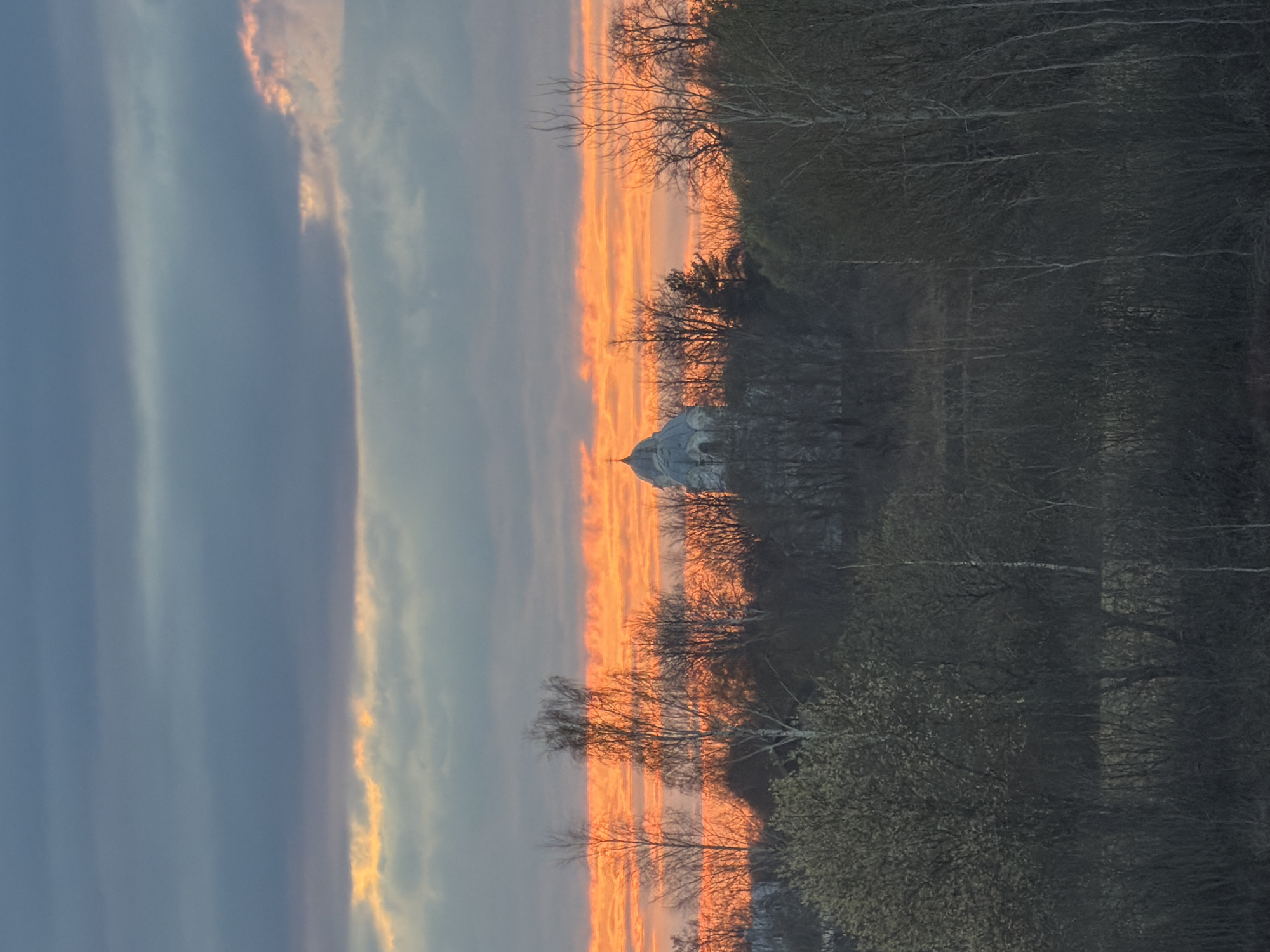 The image depicts a landscape at sunset or sunrise, featuring a classical-style building with a dome situated amidst trees. The sky is characterized by a gradient of dark blue and orange hues. The building appears to be located in a park-like setting. Specific details about the location in the city of Unknown, Unknown are not discernible from the image.