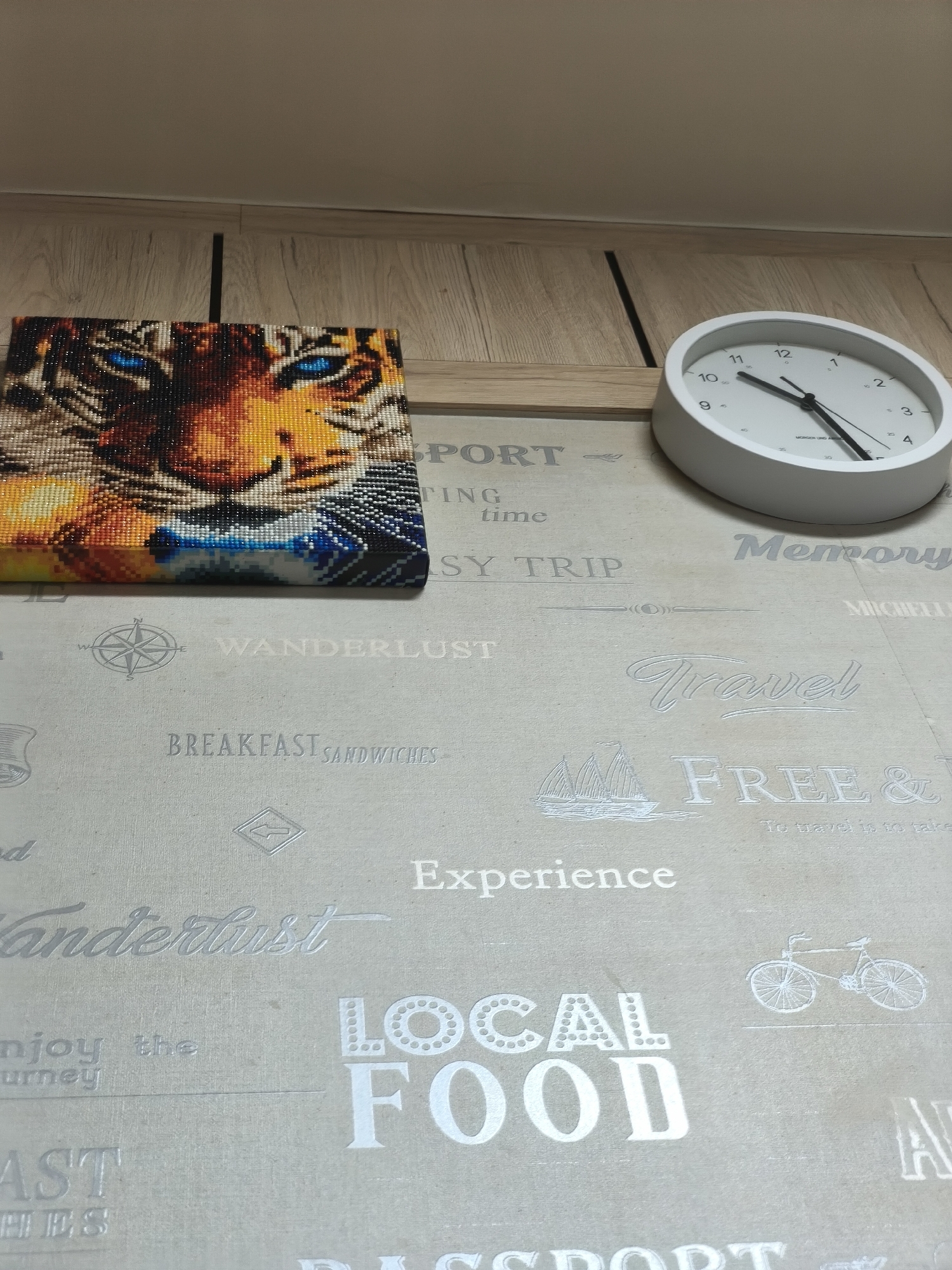 The image shows a close-up, slightly angled shot of a wall with wood paneling at the top. A circular white clock with black hands and numbers is positioned on the right side of the image, displaying approximately 10:52. To the left of the clock, a diamond art painting of a tiger's face with vivid blue eyes is displayed. Below these items, a light-colored fabric surface is visible, featuring embossed text and graphics related to travel and food. Words like "WANDERLUST," "BREAKFAST SANDWICHES," "TRAVEL," "FREE &," "EXPERIENCE," "Wanderlust," "LOCAL FOOD," and "enjoy the journey" are present. There is also a compass rose graphic, a sailboat, and a bicycle graphic. The overall scene appears to be an indoor domestic setting, likely a living room or hallway, in Asan-si, South Korea. No people are present in the image.
