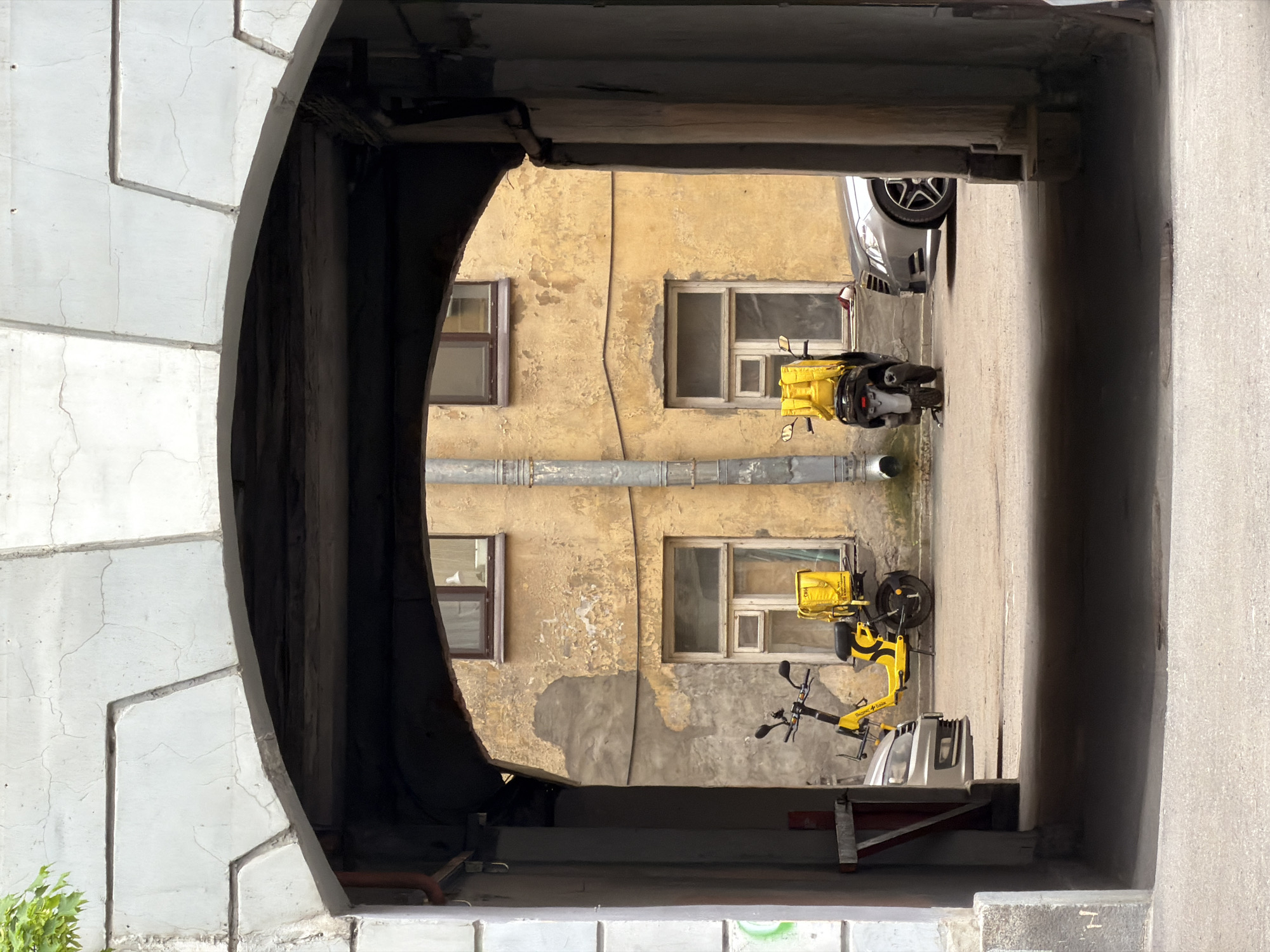 The image shows a courtyard in Saint Petersburg, Russia, viewed through an arched entrance. The courtyard features a dilapidated yellow building with windows and peeling paint. Two bright yellow delivery vehicles with large bags are parked in the courtyard. A portion of a car is visible near the end of the passageway. The arched entrance is made of grey stone or concrete with visible cracks and weathering.