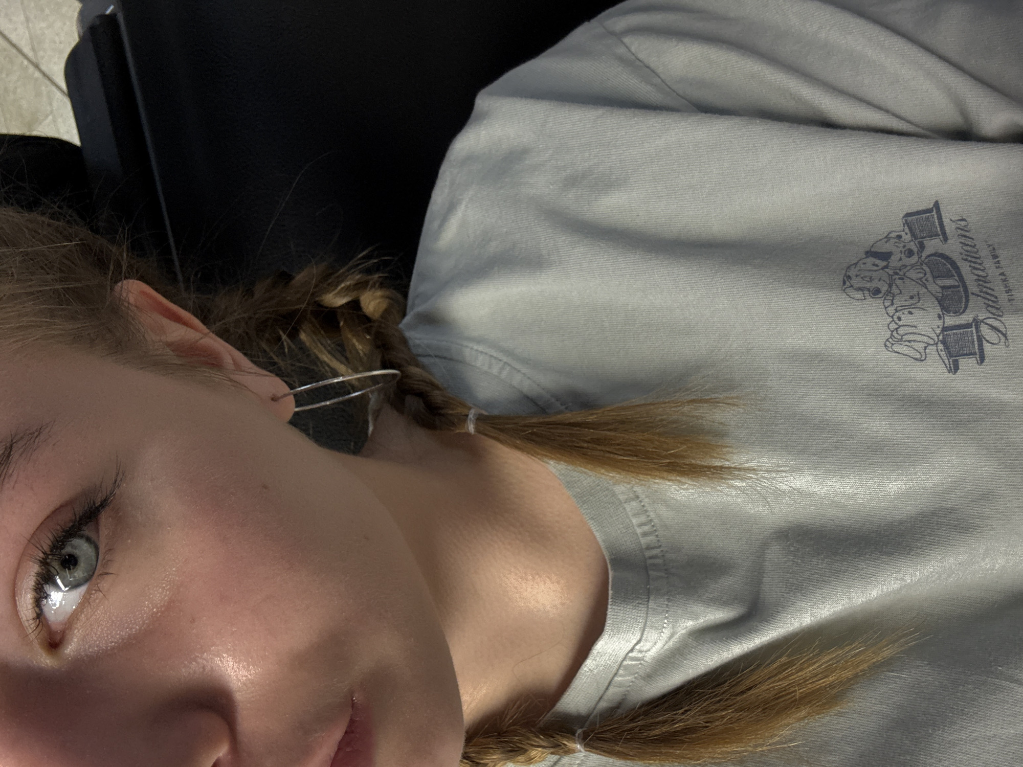 A close-up, slightly angled photograph shows a young individual with braided blonde hair and a light grey shirt featuring a "Dalmatians" graphic with two spools of thread. The person is wearing a hoop earring in their left ear. The background is out of focus, showing a black object, possibly part of a chair or other furniture. There is no discernible information within the image that would place it in Sochi, Russia.