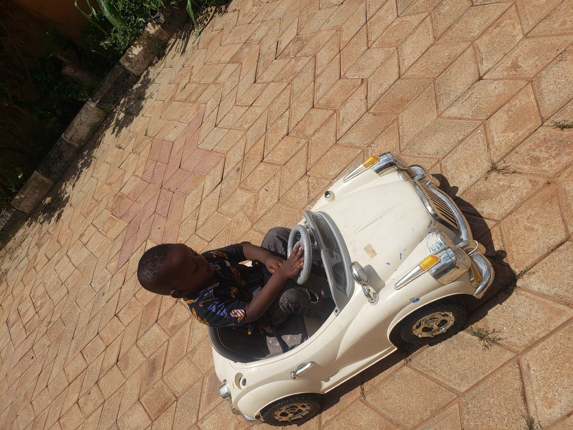 A young child, appearing to be a toddler, is seated in a white toy car on a patterned brick patio. The child is wearing a dark shirt with a colorful pattern and dark pants. Their hands are resting on the toy car's steering wheel, and they are looking down. The toy car is a retro-style convertible with silver accents, including headlights and a grille. The patio is paved with reddish-brown bricks laid in a diamond pattern. To the right of the patio, there is vegetation and a low wall. The scene appears to be outdoors, likely in a residential area. The lighting suggests it is daytime, with shadows cast by the child and the car onto the brickwork. This scene is located in Sabon Tasha, Nigeria.