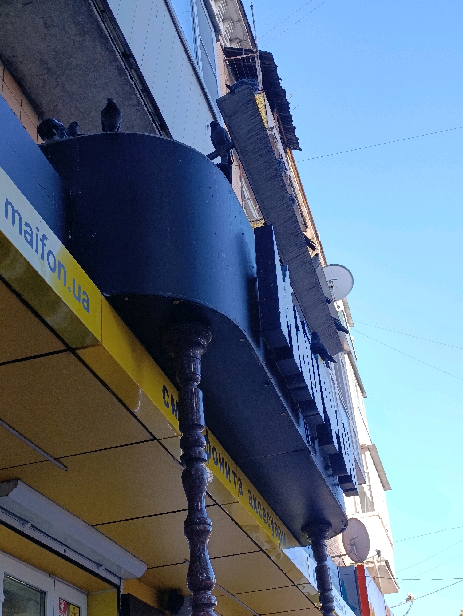 This is a low-angle, full shot photograph of the exterior of a building, likely a commercial establishment, in the city of Cherkasy, Ukraine. The sky is clear and blue, with a few thin electrical wires visible across it. The building's facade is decorated with a large, dark-colored sign with three-dimensional lettering, part of which is obscured by an overhang. Below this sign, a yellow awning runs horizontally, bearing text in blue, partially visible as "maifon.ua" and other Cyrillic characters that appear to be "СМ", followed by more Cyrillic text that is cut off. The awning is supported by decorative, dark-colored pillars with carved details. Several pigeons are perched on the ledge of the building and on the sign itself. The building's architecture suggests it is multi-story, with windows and other building elements visible above the storefront.