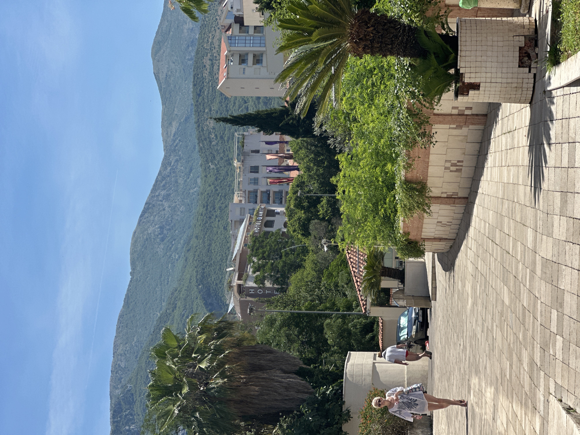 The image depicts a scene in Boreti, Montenegro, on a sunny day. In the foreground, a paved walkway with interlocking rectangular stones is visible. To the left, a woman wearing a white tunic dress with blue embroidery walks towards the viewer, carrying a patterned bag over her shoulder. Behind her, a man wearing a white t-shirt and red shorts walks away from the viewer. A dark-colored car is parked on the side of the road behind them.
The middle ground features lush green vegetation, including palm trees and shrubs. A low-lying building with a red-tiled roof is partially visible. Further back, a multi-story hotel with "HOTEL OANITA" signage is prominent, along with several colorful flags displayed vertically. Another white building with a red roof is situated to the right.
The background is dominated by a large, verdant mountain range under a clear blue sky with a faint contrail. The overall atmosphere suggests a warm, Mediterranean climate.