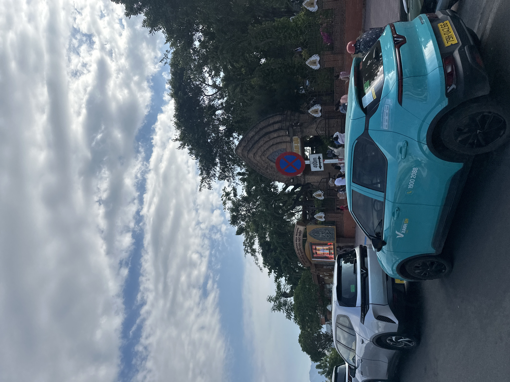 The image shows a street scene in Bac Nha Trang Ward, Vietnam. In the foreground, several cars are parked along the side of the road. The closest car is a turquoise electric vehicle labeled "Xanh SM" with the phone number 1900 2088 and the license plate "79H-042.66". Behind it are other cars, including a white SUV. In the background, a stone archway and other structures are visible, partially obscured by trees. A blue and red "no parking" sign with an arrow indicating "400m" is mounted near the archway. People can be seen walking or standing in the background. Above, the sky is filled with partly cloudy conditions. There is a sign that reads "DI TÍCH QUỐC GIA ĐẶC BIỆT".
