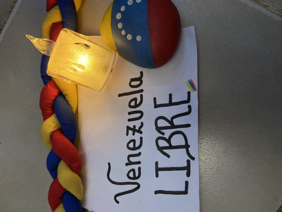 The image displays a patriotic display with a hand-painted rock in the colors of the Venezuelan flag, including the arc of stars. A braided cord in the same flag's colors sits next to the rock with a lit, decorative angel figure on top. Below, a white piece of paper shows the phrase "Venezuela LIBRE" written in black marker and the Venezuelan flag colors on the upper right. The objects are arranged on a gray surface.