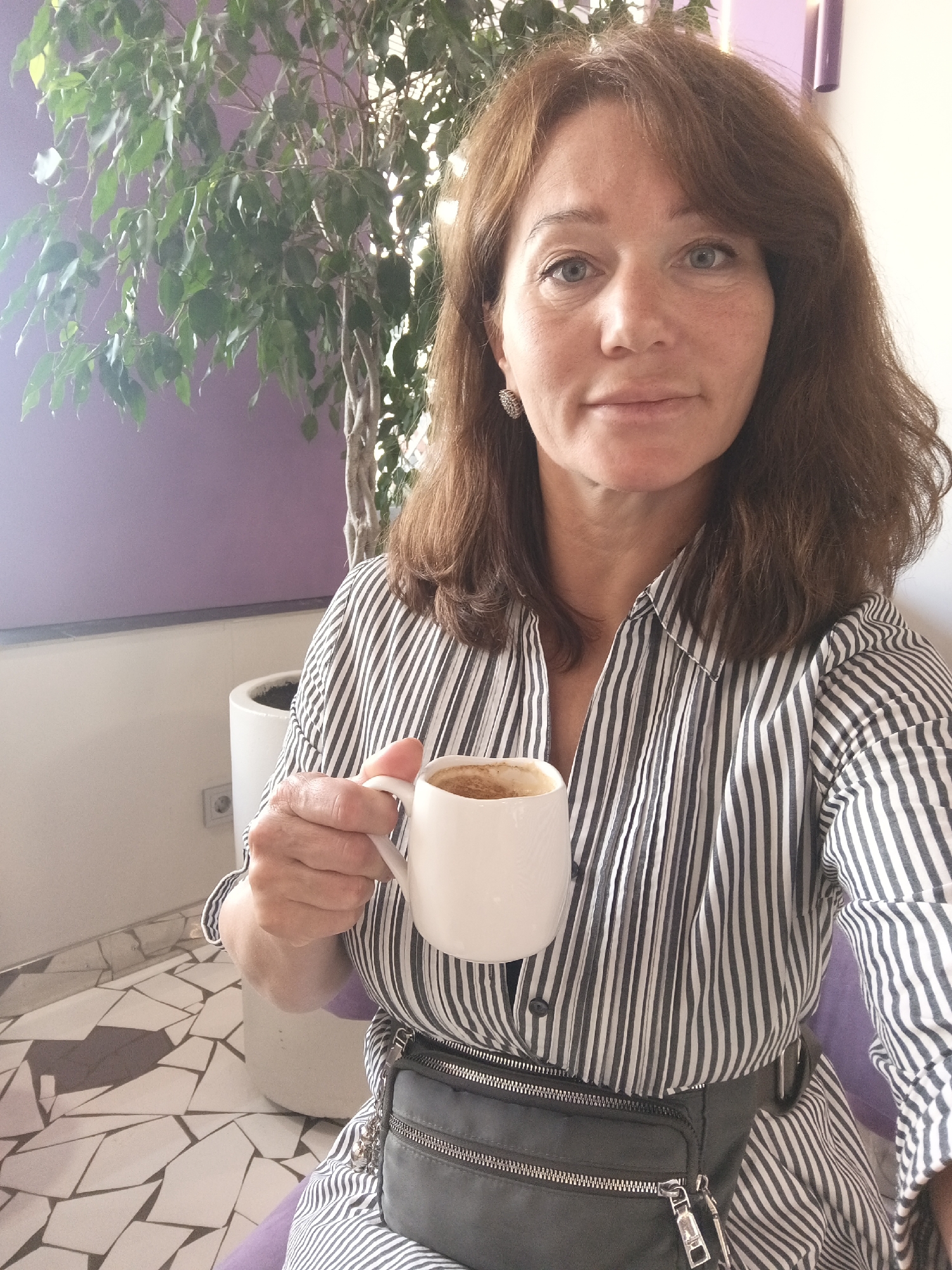A woman with shoulder-length brown hair and blue eyes is holding a white mug of coffee and smiling. She is wearing a black and white striped shirt and a dark grey fanny pack. She is sitting in front of a large plant and a purple wall. The floor is tiled with a mosaic pattern of white and grey geometric shapes. The lighting suggests it is daytime.