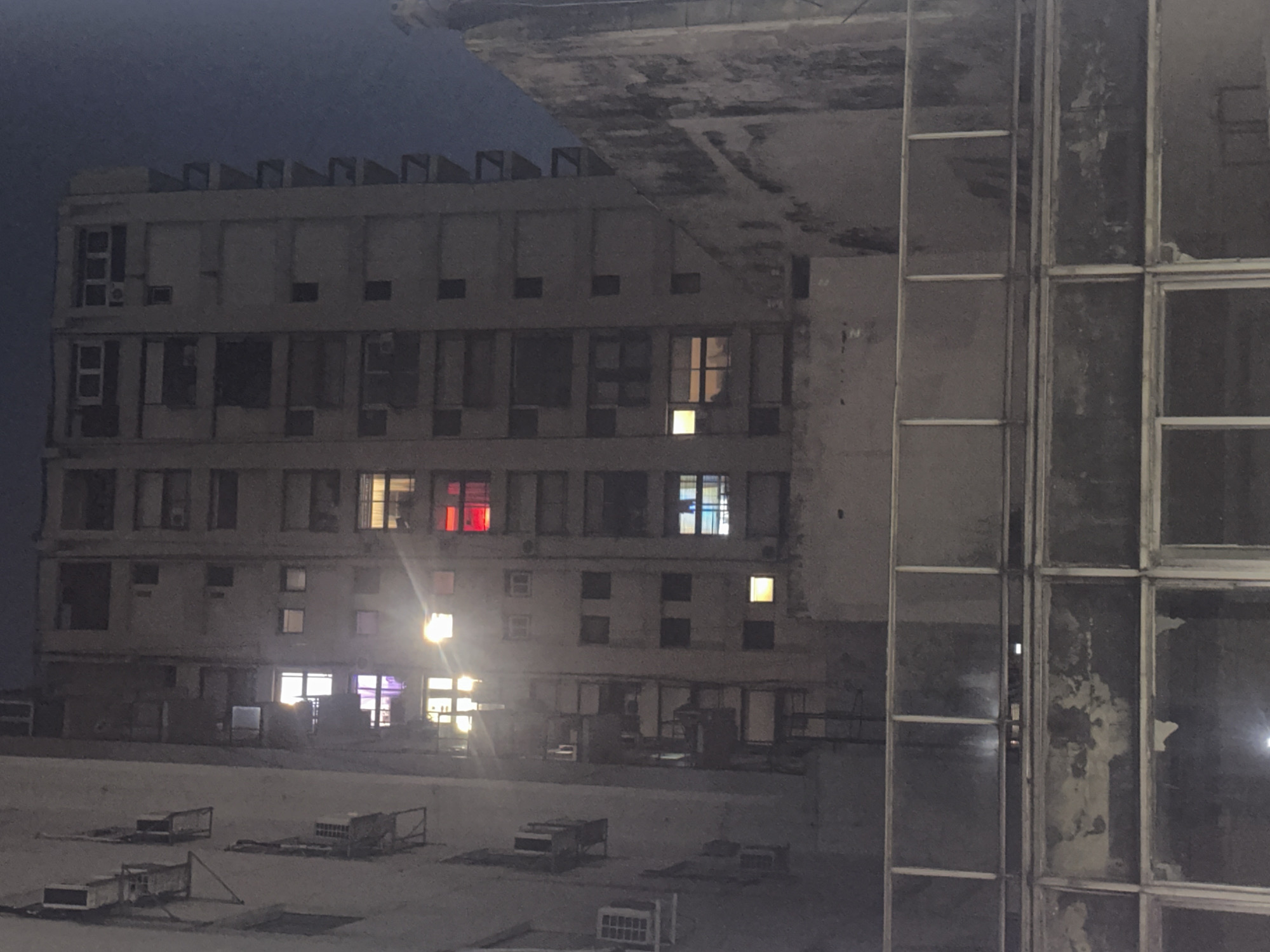 The image captures a nocturnal urban scene in Buenos Aires, Argentina, featuring a large, multi-story concrete building under a dark blue sky. The building's architecture suggests a utilitarian or possibly Brutalist design, characterized by a flat roofline with a distinctive jagged or serrated edge. Its facade is perforated by numerous rectangular windows. Many windows are dark, but several glow with artificial light, some brightly in white or yellow, and others distinctly in red on an upper floor and blue/purple on another, creating strong lens flares on the lower levels. Small air conditioning units are visible beneath some of these windows.
To the right and in the foreground, another section of the complex features large, multi-paned windows with metal frames. These windows appear dirty and reflective, with visible grime and deterioration obscuring the view into the interior, though vague shapes can be discerned behind them.
The ground level in front of the main building, or potentially a large rooftop area, is a flat, light-colored surface, possibly concrete or paved. Scattered across this surface are several rectangular objects, resembling old or decommissioned air conditioning units or other industrial equipment, some enclosed in wire frames.
The time of day is clearly night, as indicated by the dark sky and the reliance on artificial lighting from within the building. No people are visible, and there is no discernible activity taking place. The overall impression is one of quiet solitude, possibly hinting at an aging or underutilized urban space. No visible text is present in the image.