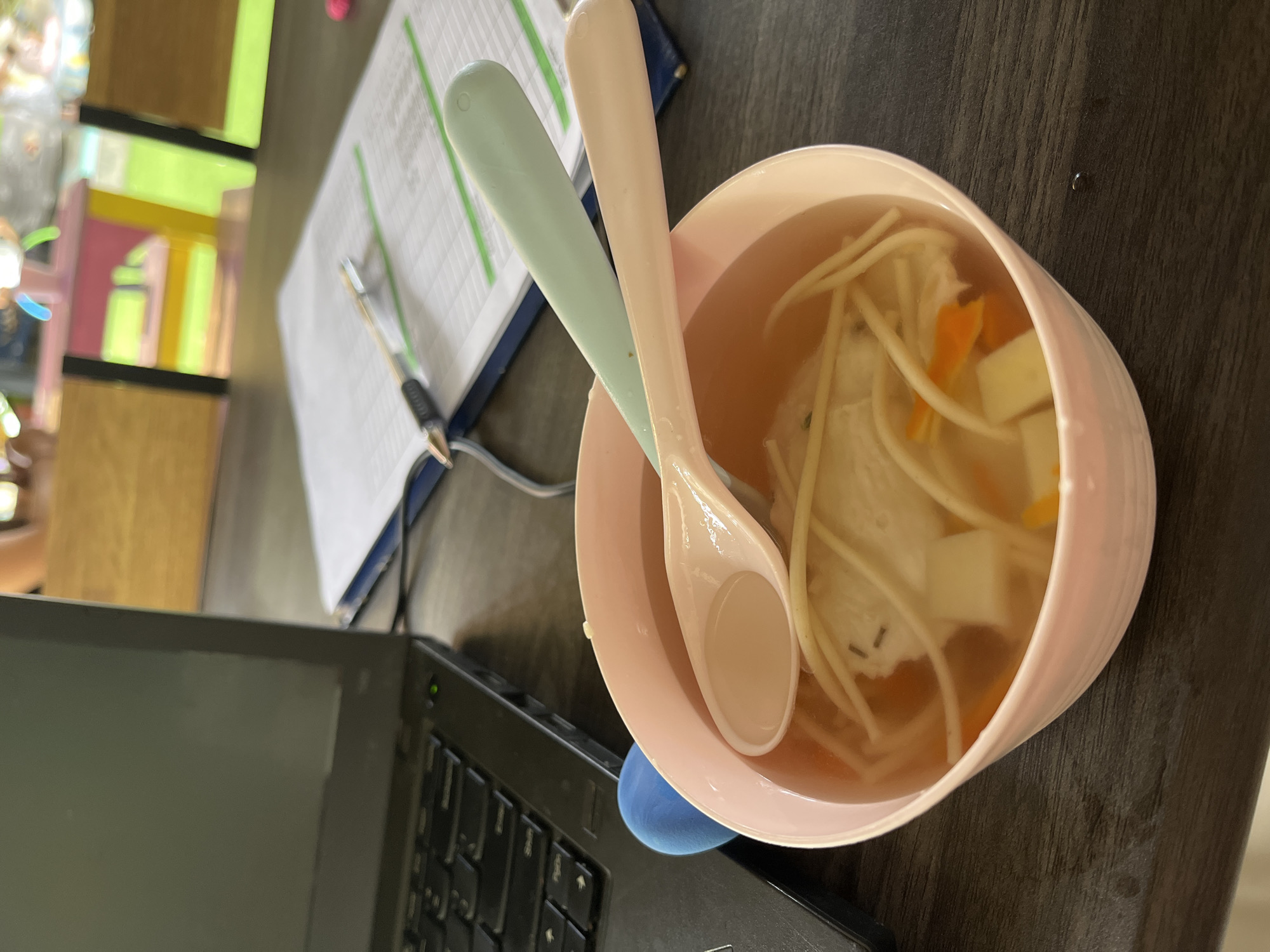A close-up, eye-level shot captures a pink bowl of soup filled with noodles, vegetables, and what appears to be a white protein, possibly chicken or tofu, placed on a dark wooden table. Two spoons, one light pink and one light teal, rest on the rim of the bowl, with the pink one submerged in the soup. To the left of the bowl is a black laptop, partially visible, with a green light illuminated on the keyboard. Behind the laptop and the bowl, a blue object, possibly a mouse or a toy, is partially visible. Further in the background, a blue folder with papers is open on the table, with a pen lying on top. The overall environment suggests a casual workspace or dining area, possibly in Kuta, Indonesia, where someone is taking a break to eat while working. The lighting is natural, indicating daytime.