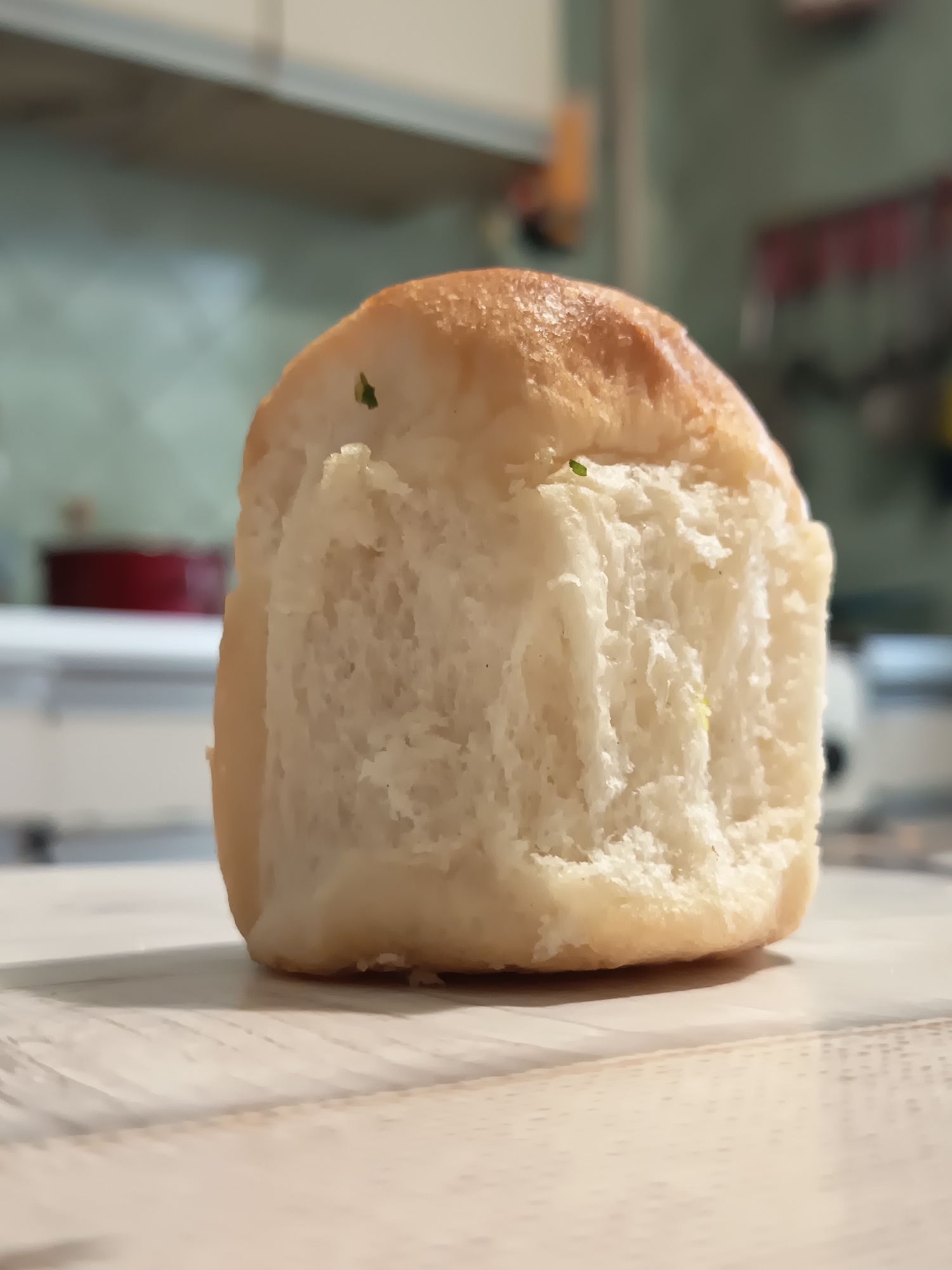 A single, freshly baked bread roll with a light golden-brown crust sits on a textured, light-colored surface, possibly a wooden table. The roll is partially torn open, revealing a soft, fluffy interior with visible air pockets. Small green specks, likely herbs, are scattered on the surface of the bread. The background is blurred, showing a kitchen environment with cabinets and appliances. The scene is brightly lit, suggesting it was photographed indoors during the day. There are no people or animals present in the image. This image does not depict any elements that would indicate its location within the city of Izmail, Ukraine.