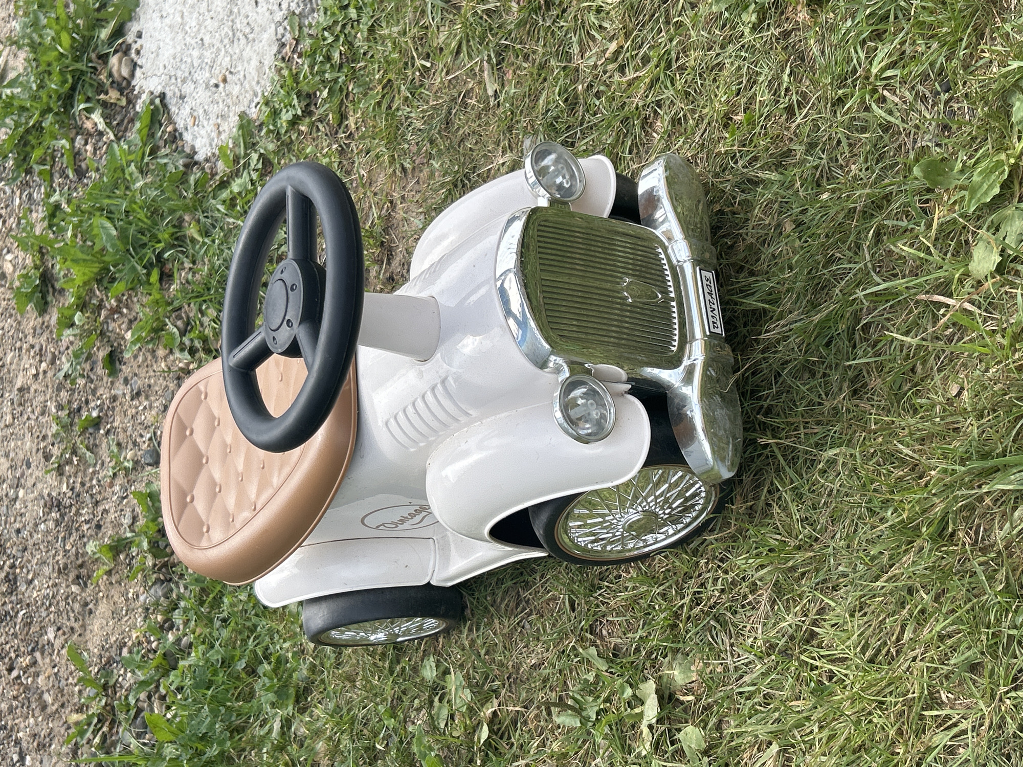 A white ride-on toy car designed to resemble a vintage automobile is parked on a grassy area with a gravel path in the background. The car has a brown, diamond-patterned seat and a black steering wheel. It features chrome accents on the grille, headlights, and wheels, which have thin spokes. A license plate on the front bumper reads "VINTAGE". The scene is outdoors, with green grass and scattered weeds. The location in the city of Chaikovsky, Russia is not discernible from the image. There are no people or actions depicted.