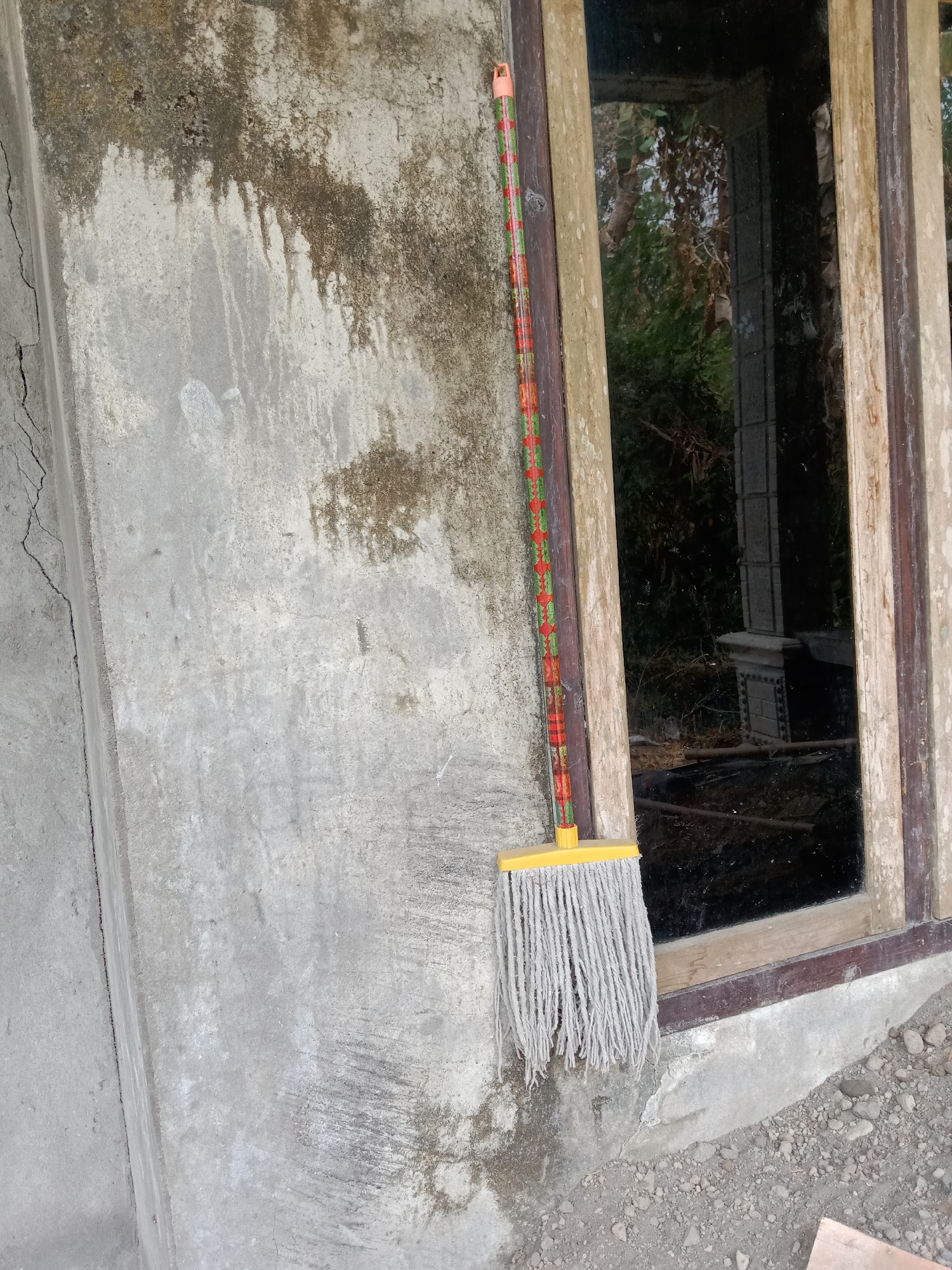A string mop with a yellow plastic head and a red, green, and orange striped handle leans against a rough, mottled concrete wall. The mop is positioned to the left of a dark-paned window with wooden trim. The window reflects trees and foliage. The ground in front of the window and wall is covered with gravel and small stones. The overall scene suggests an outdoor or unfinished area, possibly in a rural or less developed setting. The image is taken from a slightly upward angle.
