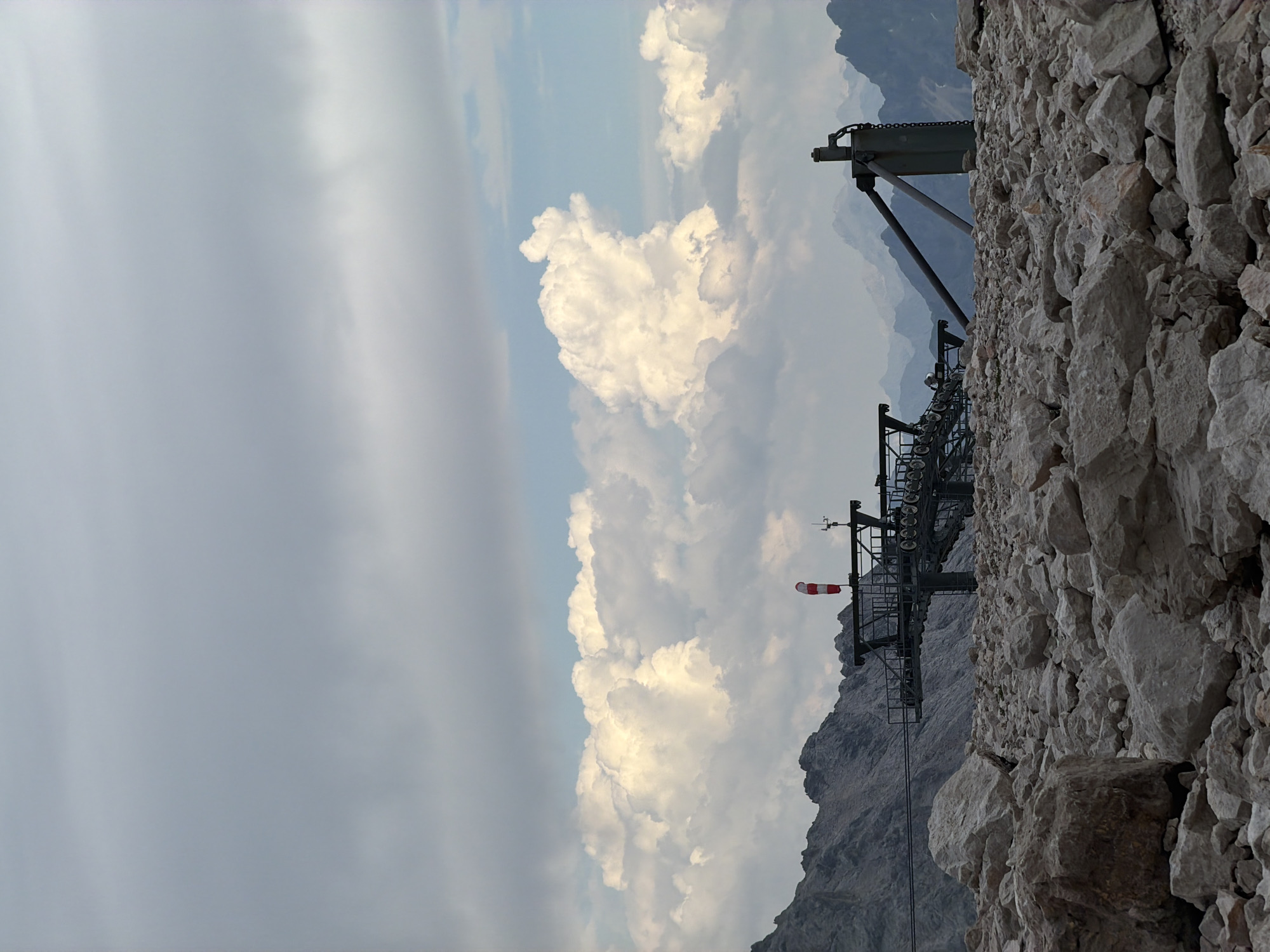 This media file depicts a mountain scene featuring a ski lift apparatus against a backdrop of dramatic clouds and rocky terrain. The ski lift, a prominent object, is seen from a low angle, with its metal structure and pulleys clearly visible. It appears to be situated on a steep, rocky mountainside, suggesting a high-altitude location. There are no people visible in the image.
The weather appears to be dynamic, with large, fluffy white clouds dominating the sky, interspersed with areas of lighter gray and blue. One of the clouds, in the mid-ground, is particularly voluminous and illuminated by sunlight, creating a striking contrast with the darker, overcast portions of the sky. A windsock, with red and white stripes, is attached to the ski lift structure, indicating wind conditions.
The time of day is difficult to pinpoint precisely, but the lighting suggests it could be late afternoon or early evening, given the soft illumination of the clouds. The terrain is characterized by rough, grey rocks, typical of a mountainous environment. In the distance, hazy mountain peaks are visible, fading into the atmospheric perspective.
The overall mood evoked by the image is one of grandeur and perhaps a touch of isolation, with the vastness of the sky and mountains contrasting with the man-made structure of the ski lift. The location context of Garmisch-Partenkirchen, Germany, implies the potential presence of ski resorts and alpine activities, though none are directly depicted. There is no visible text in the image.