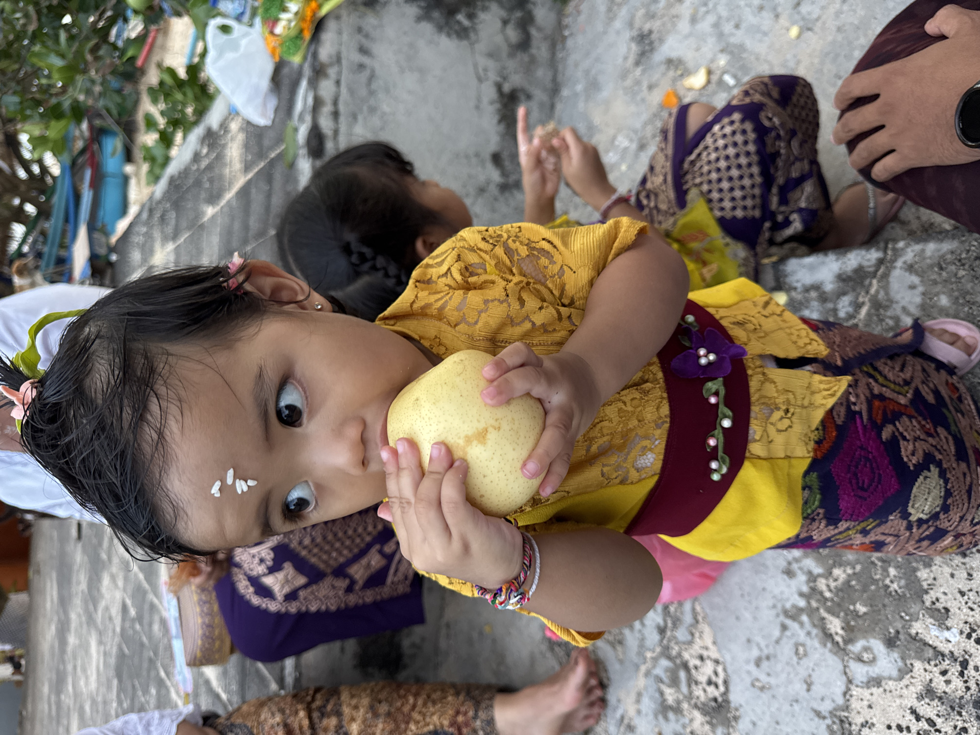 A young child in traditional Balinese attire, with white markings on their forehead, is seated on a stone surface in Kuta, Indonesia, clutching a yellow pear. The child wears a yellow lace-like top with a maroon sash adorned with purple flowers and a multi-colored patterned skirt. Beside the child, another person with braided hair is seated, while a pair of adult hands and feet are also visible in the scene. The stone surface is gray and marked with lines.