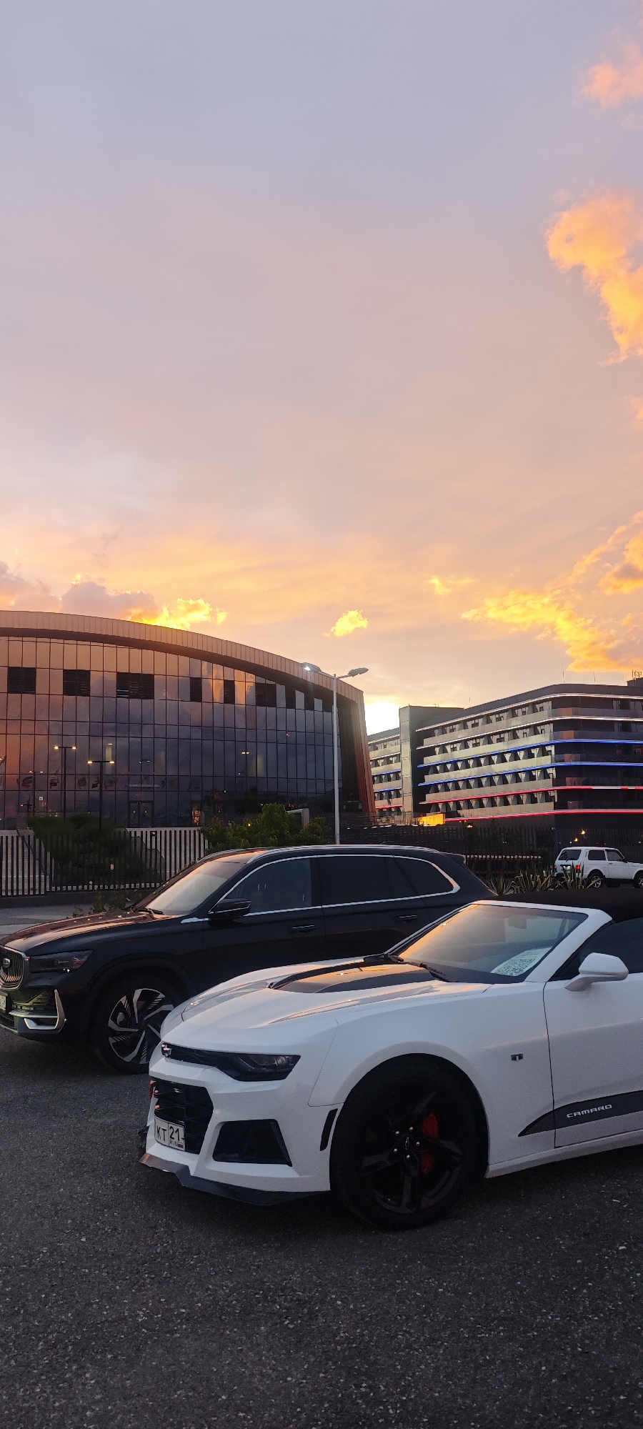 The image captures a scene in the city of Sirius, Russia, at sunset. In the foreground, a white Chevrolet Camaro convertible with black wheels and a black racing stripe along the side is parked on an asphalt surface. A black SUV, possibly a Volvo XC90, is parked behind it. The Camaro has a Russian license plate starting with "KT 21". In the background, two buildings are visible. The building to the left has a modern, curved design with reflective glass. The building on the right is a multistory structure with illuminated red and blue stripes along its facade. A white SUV is parked in front of this building. The sky is filled with clouds illuminated by the setting sun, creating an orange and pink glow. The overall environment suggests an urban setting with contemporary architecture.