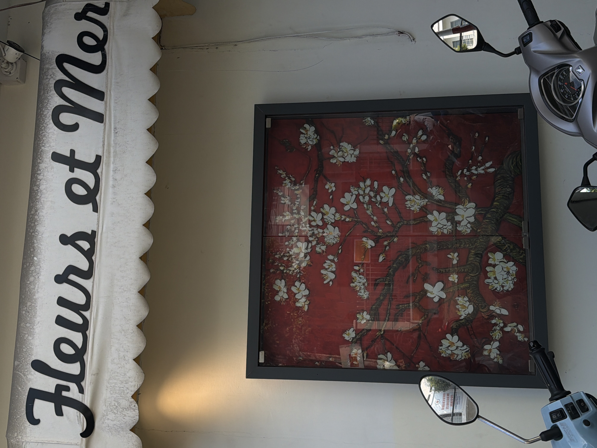 An outdoor scene in Nha Trang Ward, Vietnam, shows a close-up of a building facade and two motorcycles. Above, a white awning with a scalloped edge displays the black, stylized script "Fleurs et Mer" which translates to "Flowers and Sea". Below the awning, a framed artwork features a red background with a depiction of a flowering tree, possibly plum blossoms. The artwork is a reproduction of Vincent van Gogh's "Almond Blossoms." In the foreground, the handlebars and mirrors of two motorcycles are visible. The motorcycle on the left is light blue and the one on the right is silver. The overall setting suggests a storefront or cafe.