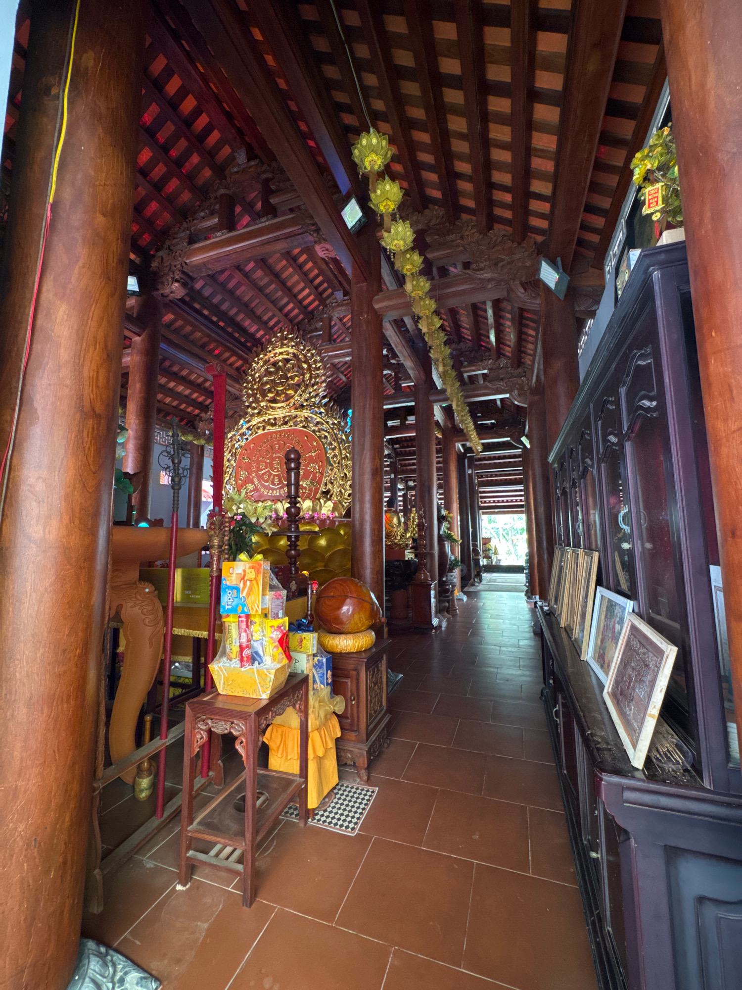 This image shows the interior of a Buddhist temple in Nha Trang, Vietnam. The temple features intricate wooden architecture with large, carved pillars supporting a high, tiled roof. In the foreground, a wooden offering table is laden with gift baskets containing snacks and drinks. Behind the table, a large, golden Buddha statue is visible, adorned with red and gold ornamentation. To the right of the Buddha, a tall, dark wooden cabinet displays framed pictures and religious artifacts. The floor is tiled in a reddish-brown color, and a walkway extends into the distance, lined with more wooden structures and leading to an open doorway. The lighting is a mix of natural light coming from the doorway and artificial lighting, creating a warm and somber atmosphere.