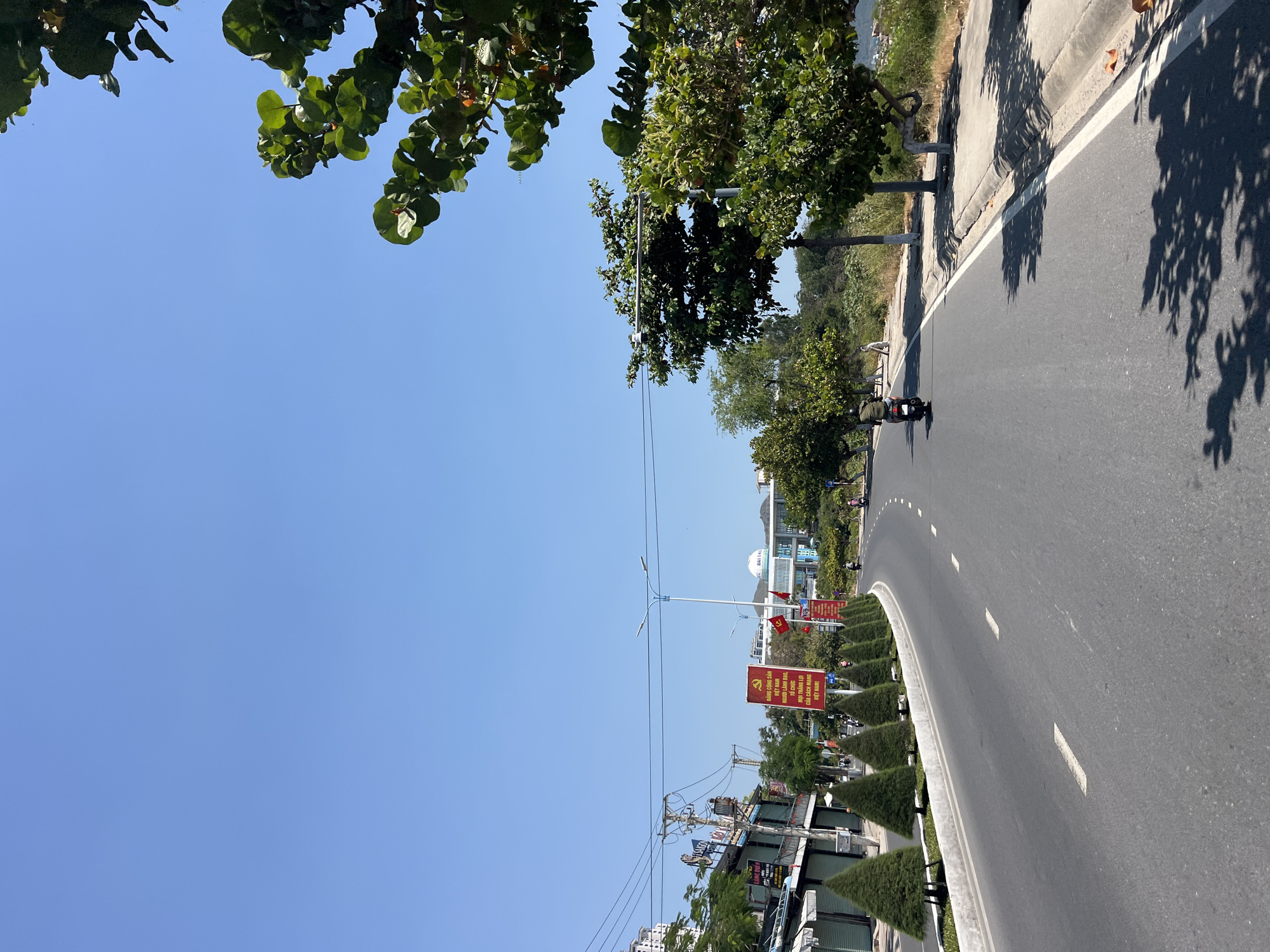 The image depicts a street scene in what appears to be Bắc Nha Trang, Vietnam, as indicated by the Vietnamese signage and architectural style. An asphalt road curves slightly to the right of the frame, marked with intermittent white lane dividers. A motorcycle is visible on the road.
On the left side of the road, there's a series of neatly trimmed, cone-shaped bushes lining the curb. Further back, structures suggest shops and residential buildings, with overhead power lines. A prominent red banner with yellow Vietnamese text and the Communist hammer and sickle emblem stands at the roadside.
On the right, the road is bordered by a concrete curb, with trees providing shade. The sky is clear and blue.
