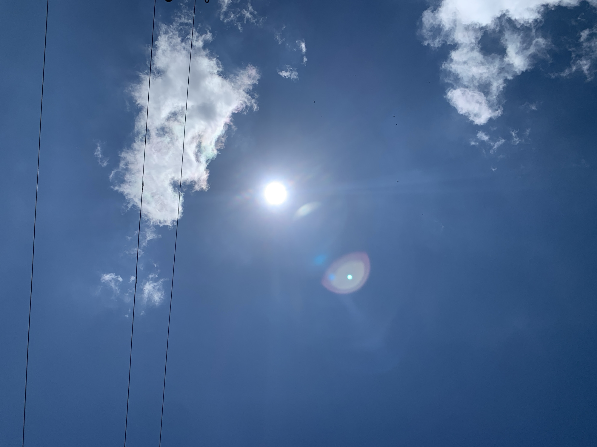 The image captures a bright, sunny sky in Kuru Karama, Nigeria. The deep blue sky is dotted with scattered white clouds. The sun is prominent, emitting a strong glare and lens flare that creates a halo effect. Several thin, dark power lines stretch horizontally across the lower portion of the frame, suggesting an urban or semi-urban setting. A few small, dark specks, possibly birds, are visible against the vast expanse of the sky. The overall impression is one of a clear and luminous day.