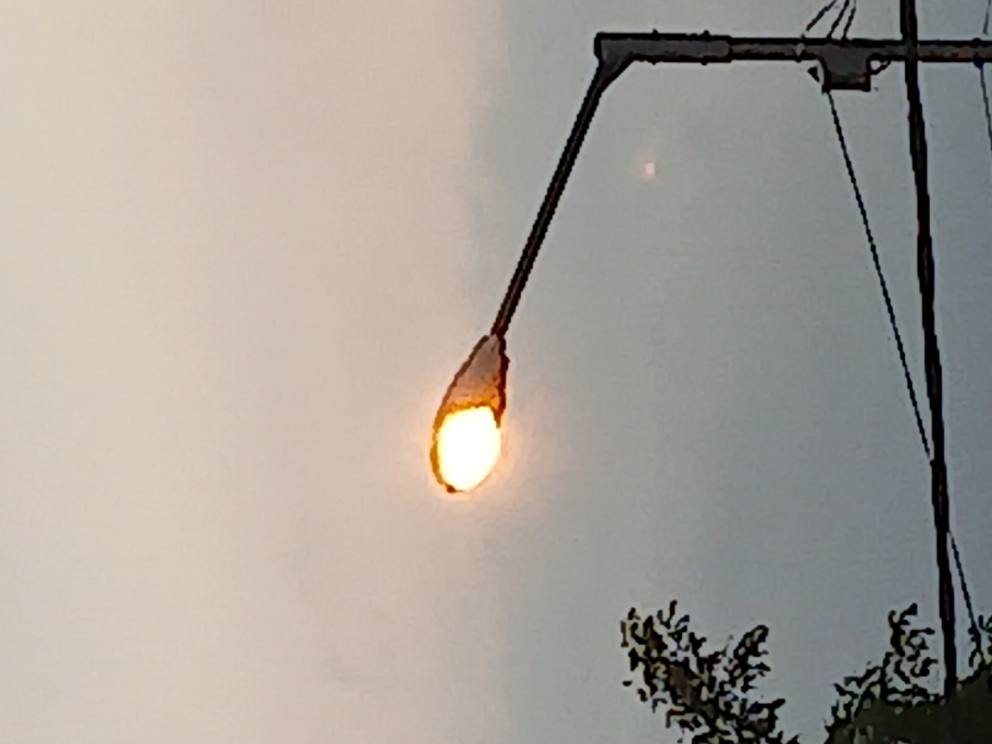 The image shows a street lamp with its light on against a cloudy sky. A portion of a tree with dark green leaves is visible on the left side, and electrical wires are strung across the lower part of the frame. The scene appears to be set in an outdoor urban or suburban environment. There are no people or any discernible actions taking place. The photograph was taken in Kademangan Kulon, Indonesia.