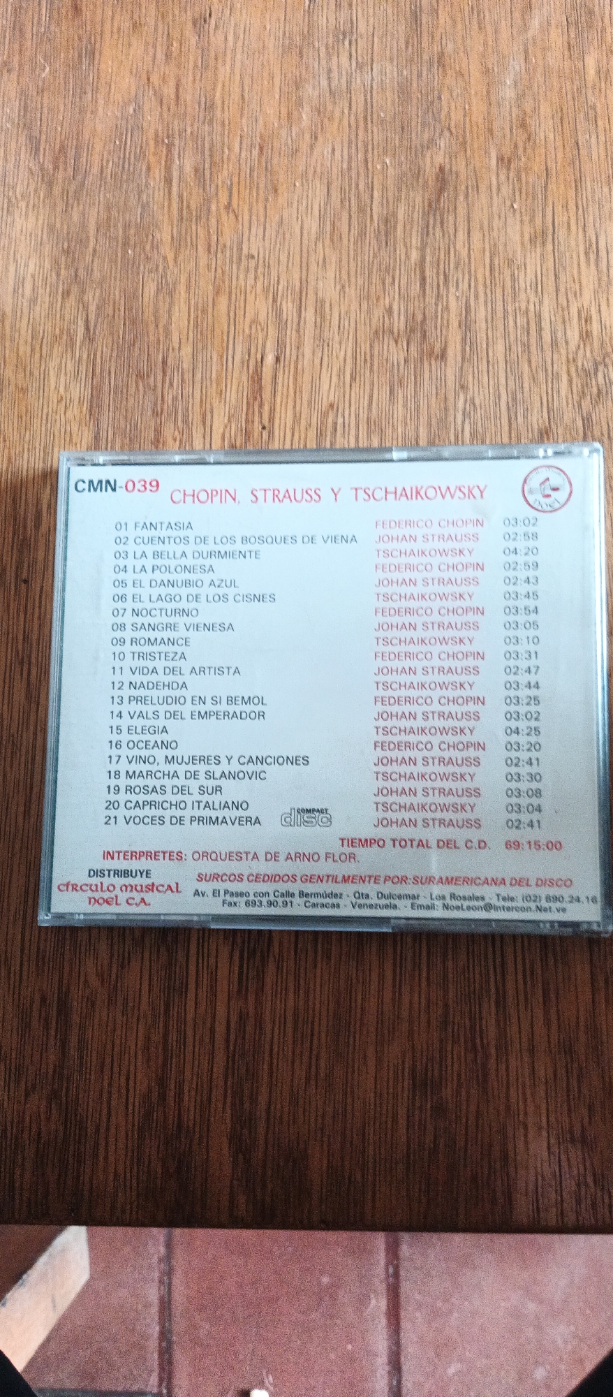 The image shows a close-up of a CD case with a tracklist. The CD case is labeled "CMN-039 CHOPIN, STRAUSS Y TSCHAIKOWSKY". The tracklist includes 21 songs with composers and durations. Below the tracklist, it indicates "INTERPRETES: ORQUESTA DE ARNO FLOR" and "DISTRIBUYE circulo musical noel C.A.". There is also contact information for "SURAMERICAN DEL DISCO" including an address in Caracas, Venezuela. The CD case is placed on a surface that appears to be made of wood. There are no people or specific environmental details beyond the CD case and the surface it rests on. The city of La Tagua, Venezuela, is not mentioned or depicted in the image.
