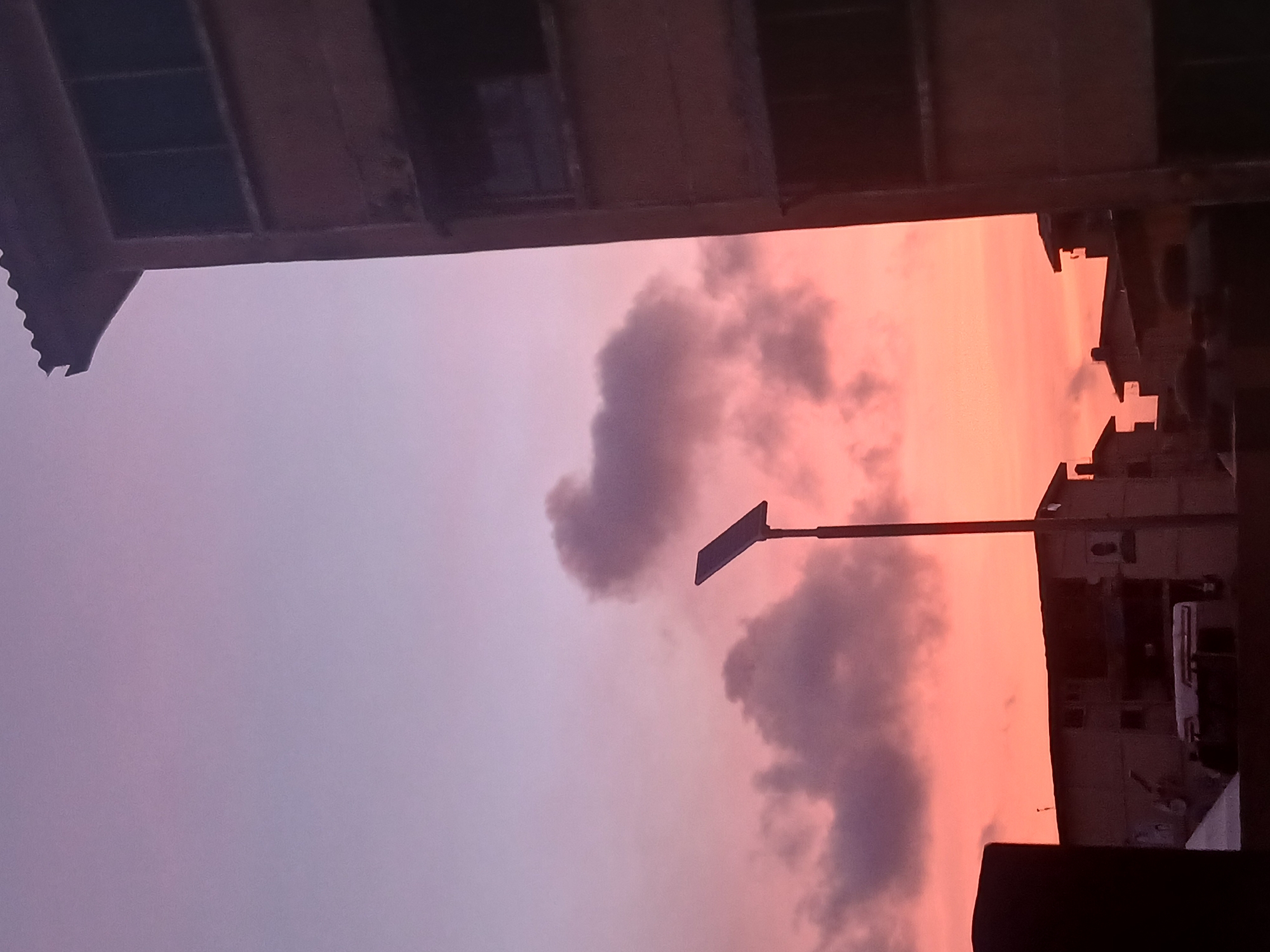The image shows a sunset view in Lagos, Nigeria, framed by the architectural structures of the city. In the foreground, parts of a building's roof or balcony are visible at the top and bottom of the frame, creating a narrow window onto the scene. The sky transitions from a pale lavender color on the left to a vibrant orange-pink hue on the right. Dark, puffy clouds are scattered across the sky. A street lamp with a rectangular panel is visible, casting a silhouette against the colorful sky. In the background, buildings are visible against the sunset, silhouetted against the vivid colors. The image captures a moment of urban beauty in Lagos.