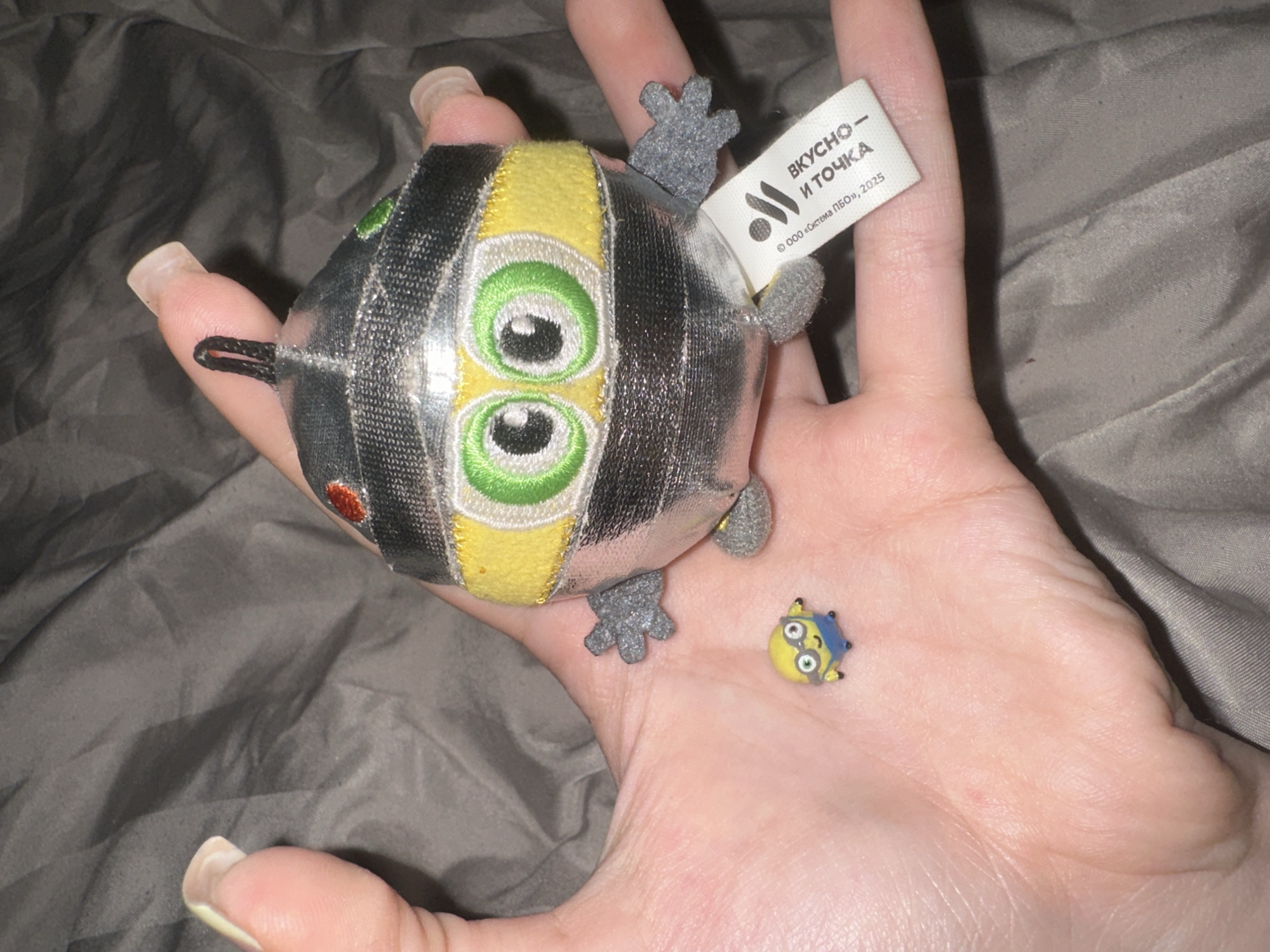 A hand is holding a small, plush toy with a silver metallic body, a yellow band around its middle, and large green eyes. The toy has gray fabric hands and feet. Attached to the toy's side is a white tag with Russian text and a logo. To the left of the plush toy, on the palm of the hand, is a miniature figurine of a yellow character with goggles and blue shorts, resembling a minion. The background is a gray, textured fabric. The image is taken indoors. It is not possible to determine the location in the city of Petropavlovsk-Kamchatsky, Russia based on the provided image.