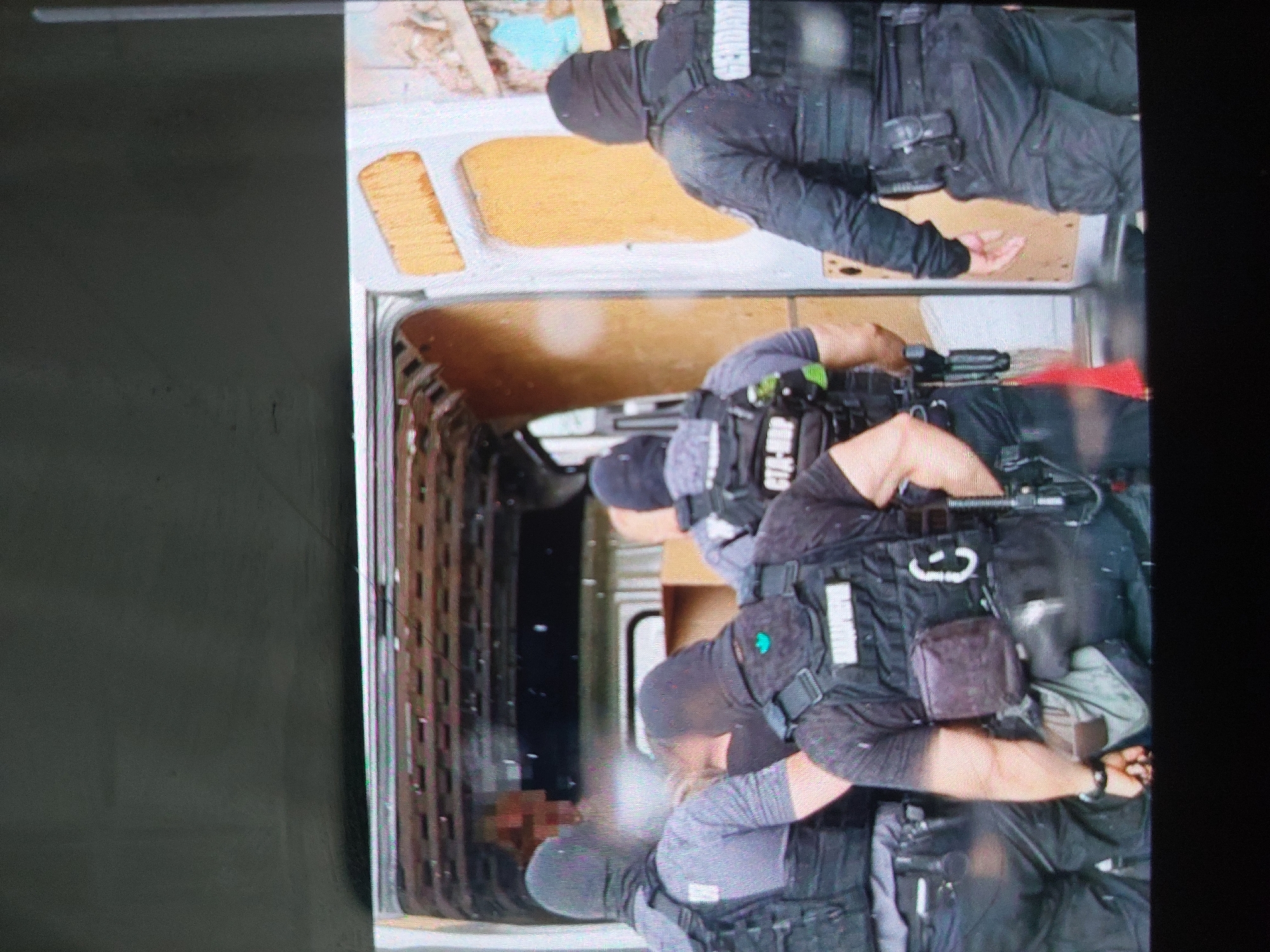 The image depicts the interior of a van, likely in the city of Tsarevo, Bulgaria, containing at least four individuals dressed in tactical gear and balaclavas. They appear to be law enforcement or military personnel, based on their uniforms and equipment, including firearms, tactical vests, and restraints. The individuals are restraining at least one person whose face has been blurred. The van’s interior is plain, with wooden panels and metal framework visible. The overall scene suggests an arrest or apprehension operation.
