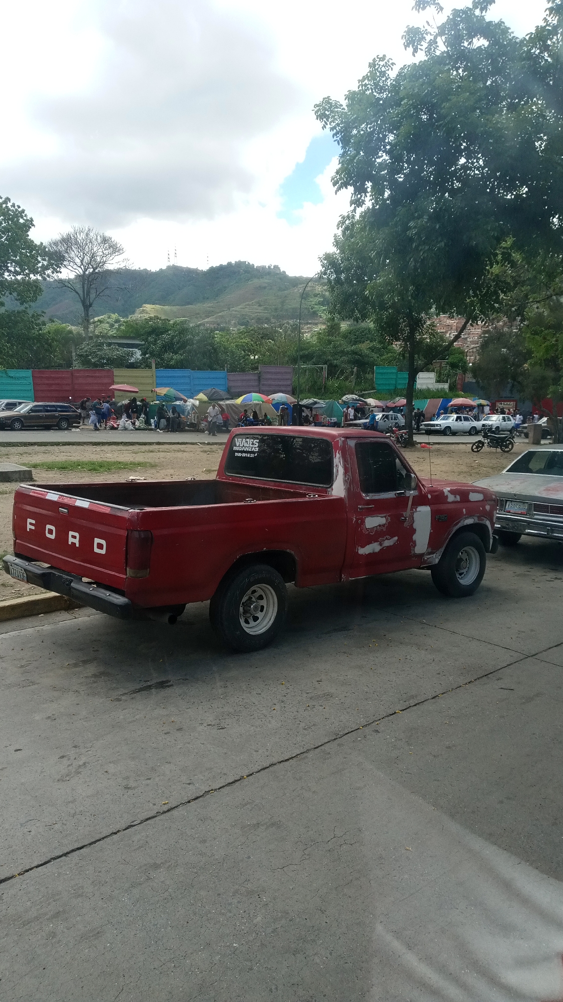 The image depicts a red Ford pickup truck parked on a paved surface. The truck shows signs of wear and tear, with faded and peeling paint. The rear of the truck has the word "FORD" in large white letters. The truck is parked near a group of tents and people, suggesting a possible encampment or gathering. In the background, there are hills or mountains. The location is unknown, but appears to be an urban area with signs of temporary habitation.