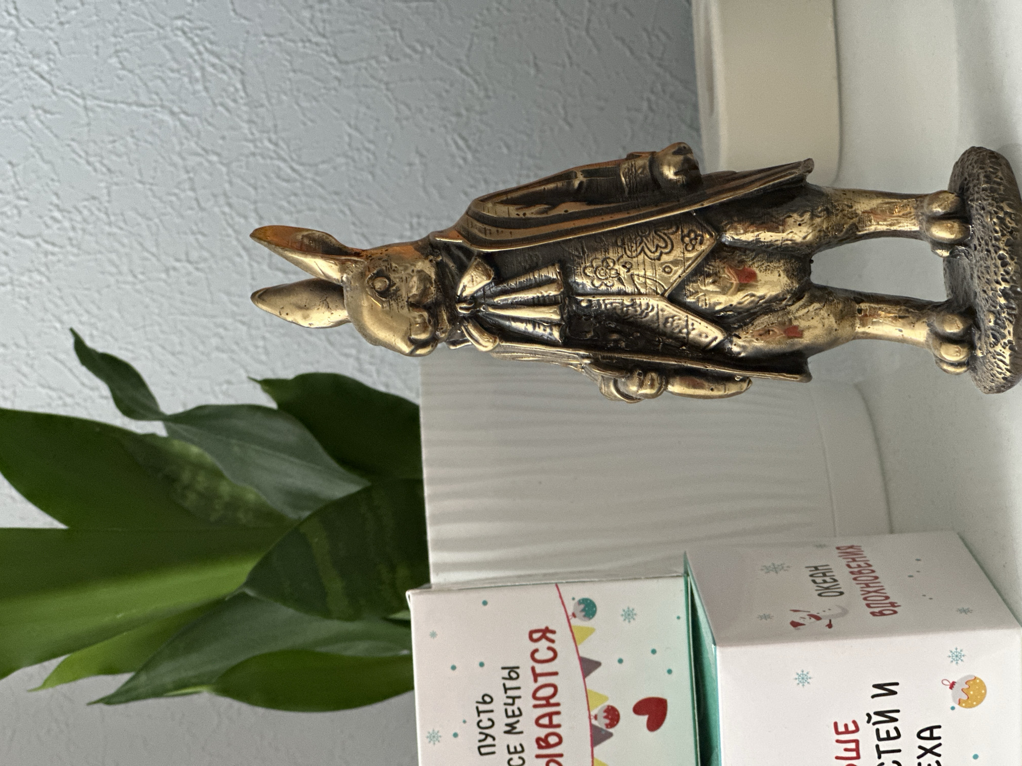 A metallic, antique-looking figurine of a rabbit dressed in formal attire stands on a textured, circular base. The rabbit is wearing a coat with intricate patterns, a vest, and a bow tie. To the left of the figurine, there are two stacked cardboard boxes. The top box is white with blue dots, snowflakes, and text that includes the word "МЕЧТЫ" (dreams). It also features a red heart and a festive garland with small ornaments. The bottom box is also white and displays snowflakes, a snowman, and Russian text, including "ОКЕАН ВДОХНОВЕНИЯ" (ocean of inspiration). Behind the boxes and the figurine, the broad, green leaves of a potted plant are visible, along with a portion of a white textured wall. The overall scene suggests an indoor setting, possibly a shelf or table, with decorative items. The location is specified as Nevinnomyssk, Russia.