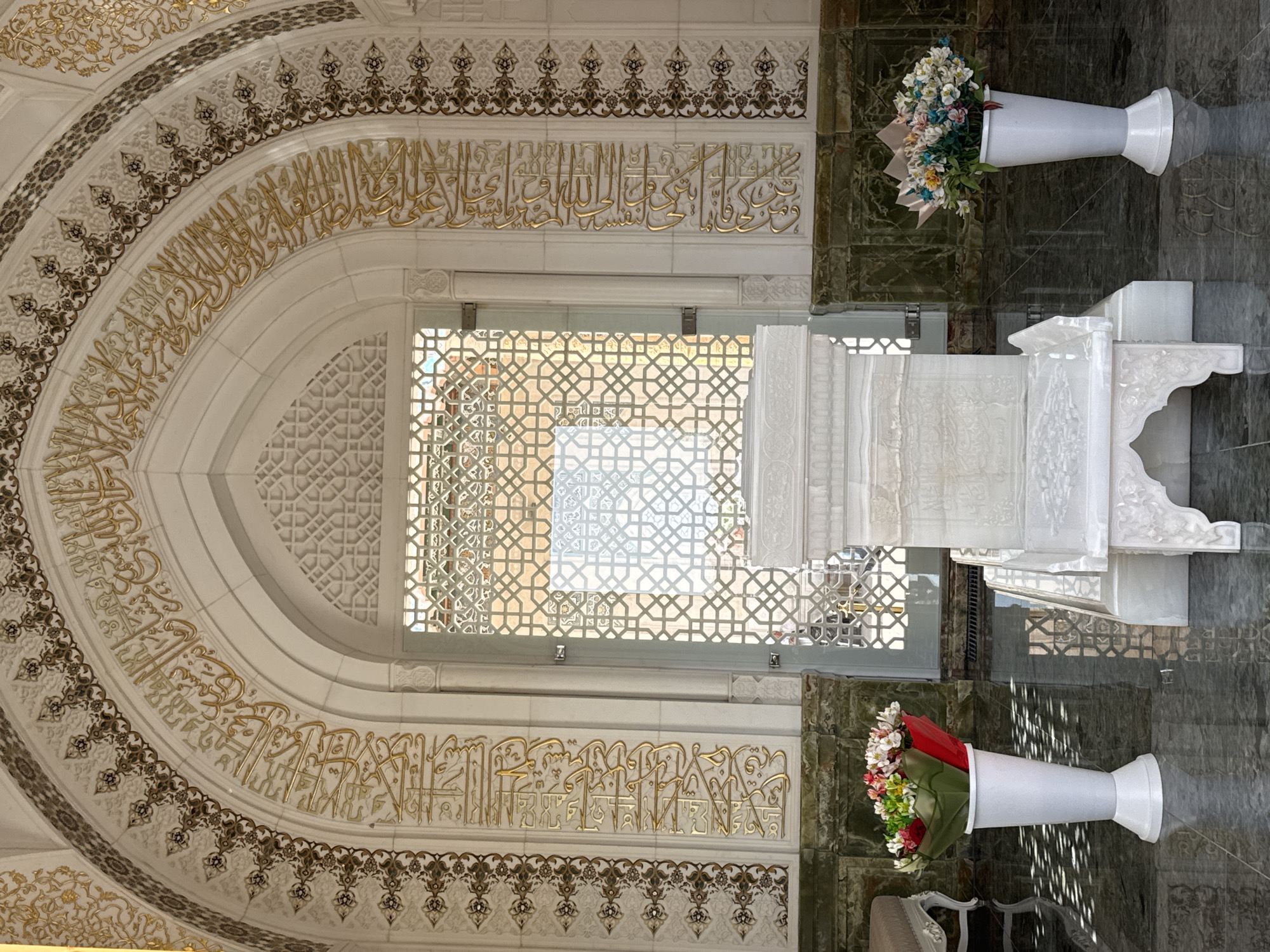 The image shows the interior of a building with Islamic architecture. An archway with decorative gold-colored Arabic calligraphy frames a window with an intricate geometric pattern. Below the archway, a similar inscription runs along the wall. Two white vases containing flowers stand on either side of a white marble structure, possibly a cenotaph or tomb. The background floor is a dark green with some brown marble pattern. The architecture and decorative elements suggest a mosque or a mausoleum. Based on the style and details, it's highly plausible that this is within the city of Samarkand City, Uzbekistan.