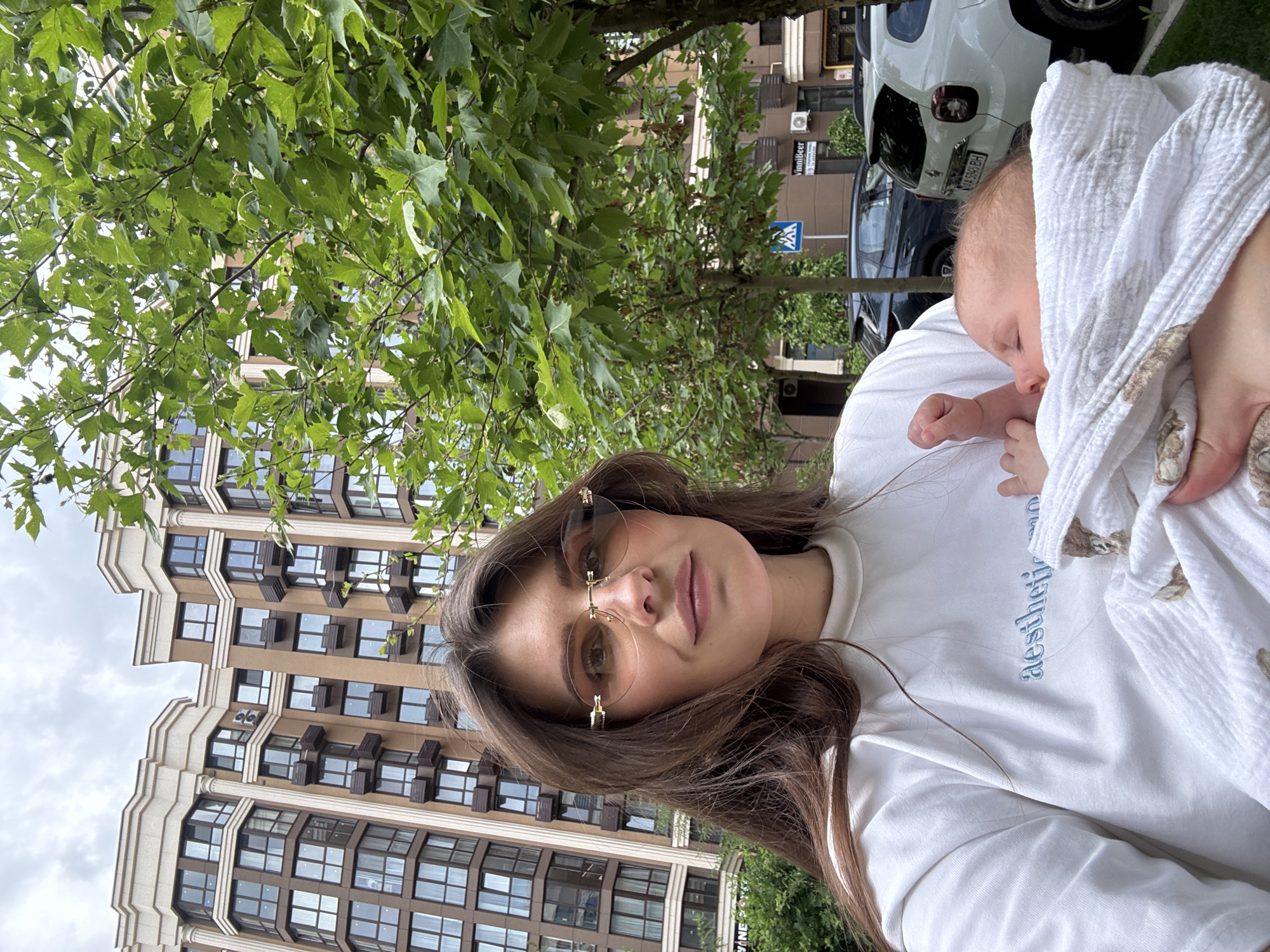 The image depicts a woman holding an infant outdoors in Sofiivska Borshchahivka, Ukraine. The woman, positioned centrally, has long brown hair and wears rimless, lightly tinted sunglasses with gold-colored frames. She is dressed in a white t-shirt with "aesthetic" embroidered in light blue text on the chest. She holds a swaddled infant wrapped in a white blanket with a faint brown pattern. The infant's head is visible, turned to the viewer's left, with eyes closed, indicating sleep. The infant's right hand is partially curled.
In the background, a multi-story residential building with a brown facade and beige architectural trim is prominent. It features numerous rectangular windows and small decorative balconies or window boxes. Two air conditioning units are visible on an upper floor. Green deciduous trees with broad leaves are situated on the right side of the frame and between the foreground subjects and the building. A street is visible beneath the trees and in front of the building's lower levels, featuring parked vehicles. A white SUV, identified by the Ukrainian license plate "KA 5965 BH," is parked on the right, with a dark-colored SUV behind it. A blue and white pedestrian crossing sign is visible near the parked cars. A commercial sign reading "VinniBeer" is visible on the building's ground floor, while another partial sign reading "WINE" is on the left. The sky is overcast with a consistent cloud cover.