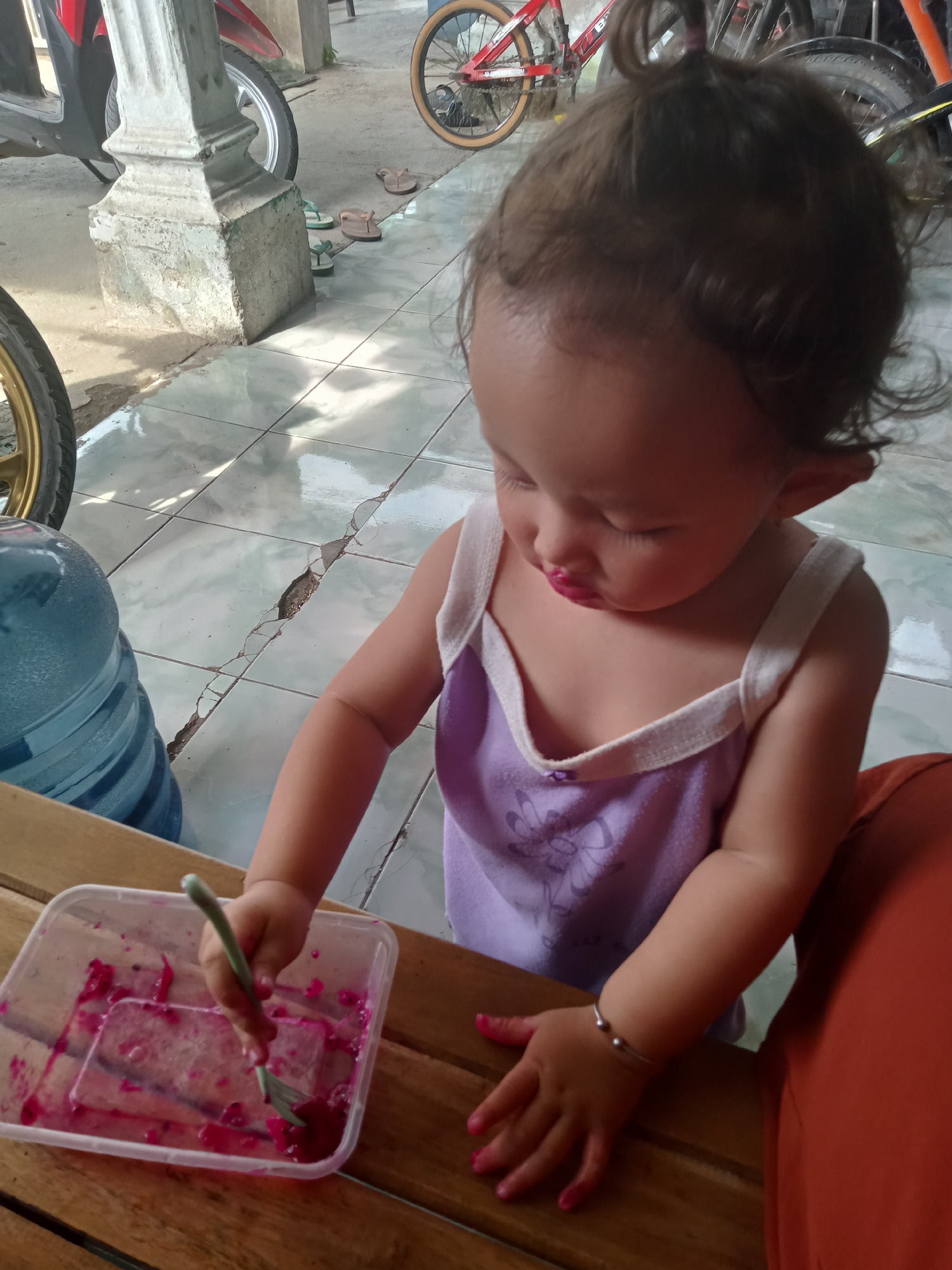 A young child, likely a toddler, is depicted in a brightly lit, semi-outdoor setting, possibly a porch or open-air area, in Bungur, Indonesia. The child has light brown hair tied up on top of their head and is wearing a light purple tank top with a white trim and a subtle graphic design. A silver bracelet with two beads is visible on their right wrist.
The child is focused on a clear plastic container placed on a wooden table, which holds a quantity of bright pink or red fruit, appearing to be dragon fruit, with some juice. They are holding a light green fork and are in the process of scooping or playing with the fruit. Their lips are stained pink/red from the fruit, and their fingernails are painted a matching vibrant pink.
In the background, elements of the environment are visible. To the left, a large blue water dispenser bottle sits on the ground next to a tire, likely from a motorcycle, which is partially visible. Further back, a concrete pillar stands on a light-colored tiled floor that shows signs of cracking. Several pairs of flip-flops are scattered near the pillar. Parts of motorcycles and bicycles, including wheels and frames, are discernible in the deeper background, suggesting a common domestic or communal space. The bright natural lighting indicates it is daytime. No visible text is present in the image.