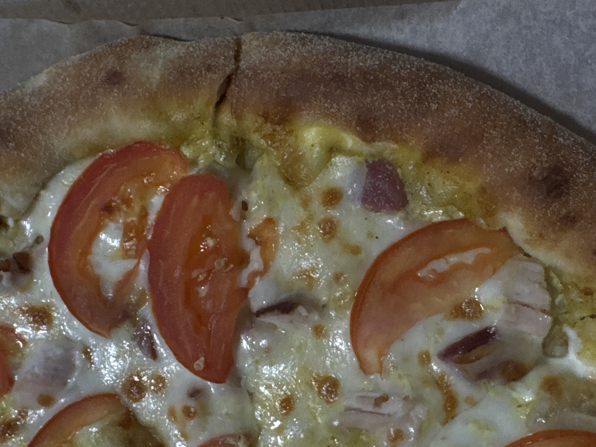 This is a close-up, overhead shot of a pizza, likely taken indoors. The pizza has a thick, golden-brown crust with a slightly bubbled texture, suggesting it's been baked. Slices of red tomato are scattered across the cheesy surface, interspersed with pieces of what appears to be red onion and possibly some meat, like ham or bacon. The cheese is melted and bubbly, with some areas appearing slightly browned. The pizza is resting in a cardboard box, indicated by the textured brown material visible in the background. There is no text visible in the image. The lighting suggests an indoor setting, possibly a kitchen or dining area, with no specific time of day or weather cues discernible. The overall impression is of a freshly prepared and appealing dish.