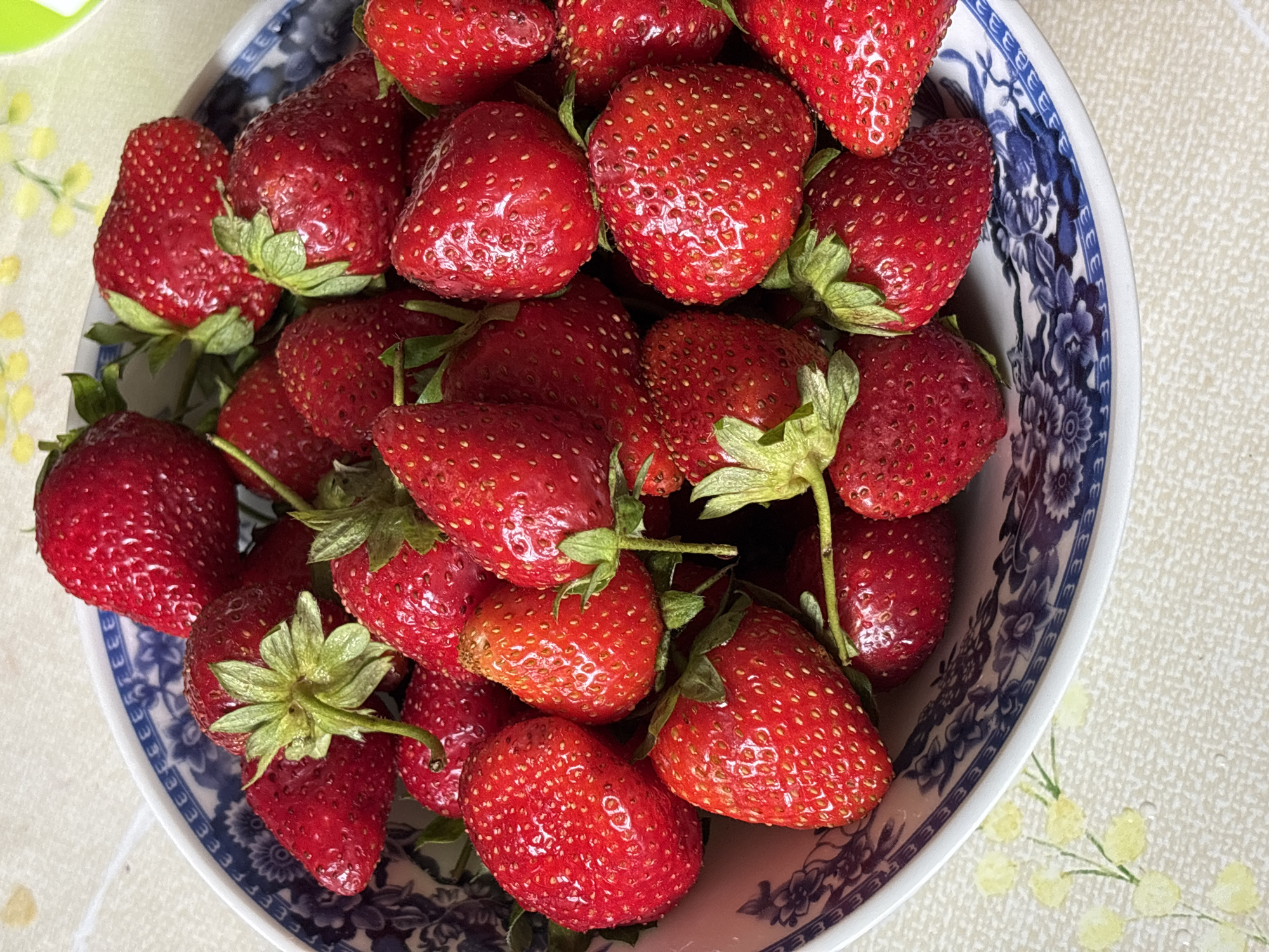 A close-up, overhead shot displays a bowl filled with ripe, red strawberries. The bowl has a white base with a blue, floral pattern around its rim. The strawberries are a vibrant red, some with visible seeds, and green stems and leaves. The bowl sits on a light-colored tablecloth with a subtle pattern of yellow flowers. The scene is well-lit, highlighting the texture and color of the fruit. The image does not depict any people or specific landmarks that would indicate a location in Новый Кинер, Russia.