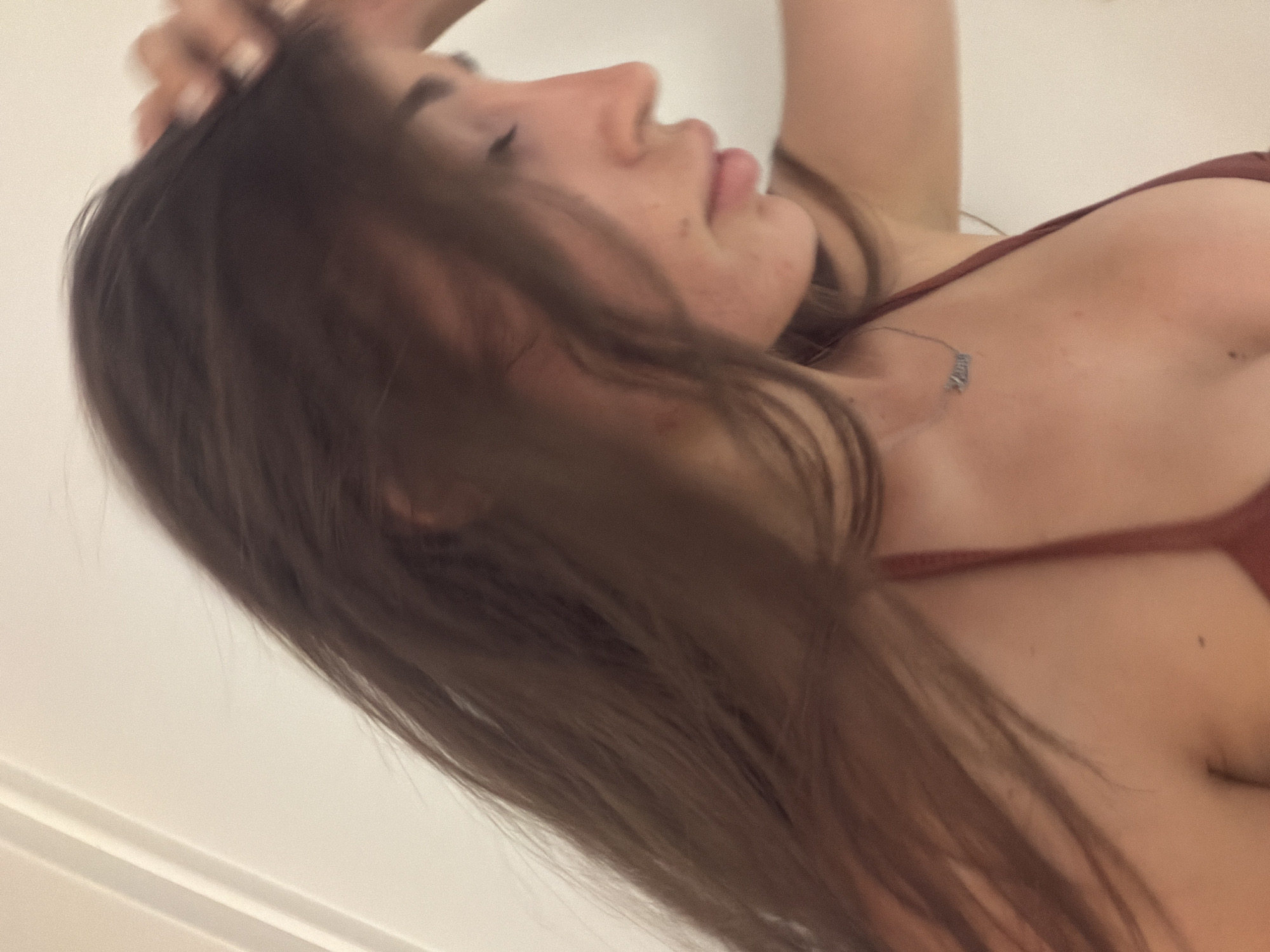 A young woman with long, dark brown hair is captured in a close-up, profile view against a plain, light-colored background, possibly an indoor wall or door. She is wearing a dark reddish-brown halter-neck top, likely a bikini or bra, with a delicate silver necklace featuring a word pendant. Her eyes are closed, and her right hand is raised, gently touching her hair near the crown of her head. The lighting suggests it could be natural light coming from the side, creating subtle shadows on her face. The overall tone of the image is casual and personal, with a slightly soft focus that emphasizes her features. The location is not discernible from the image provided.